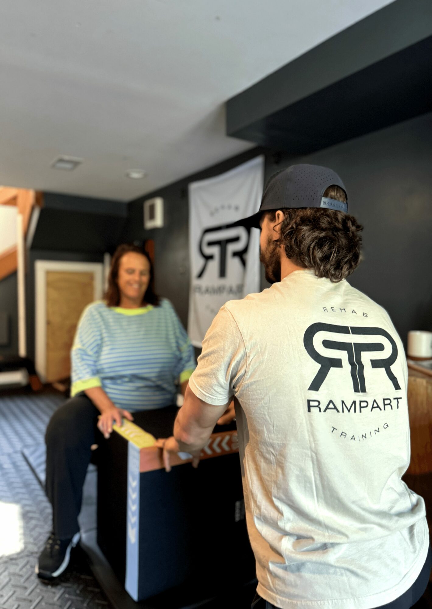 Two people talking indoors, one wearing a cap and a shirt with a logo, the other woman in a striped shirt, in front of a banner.