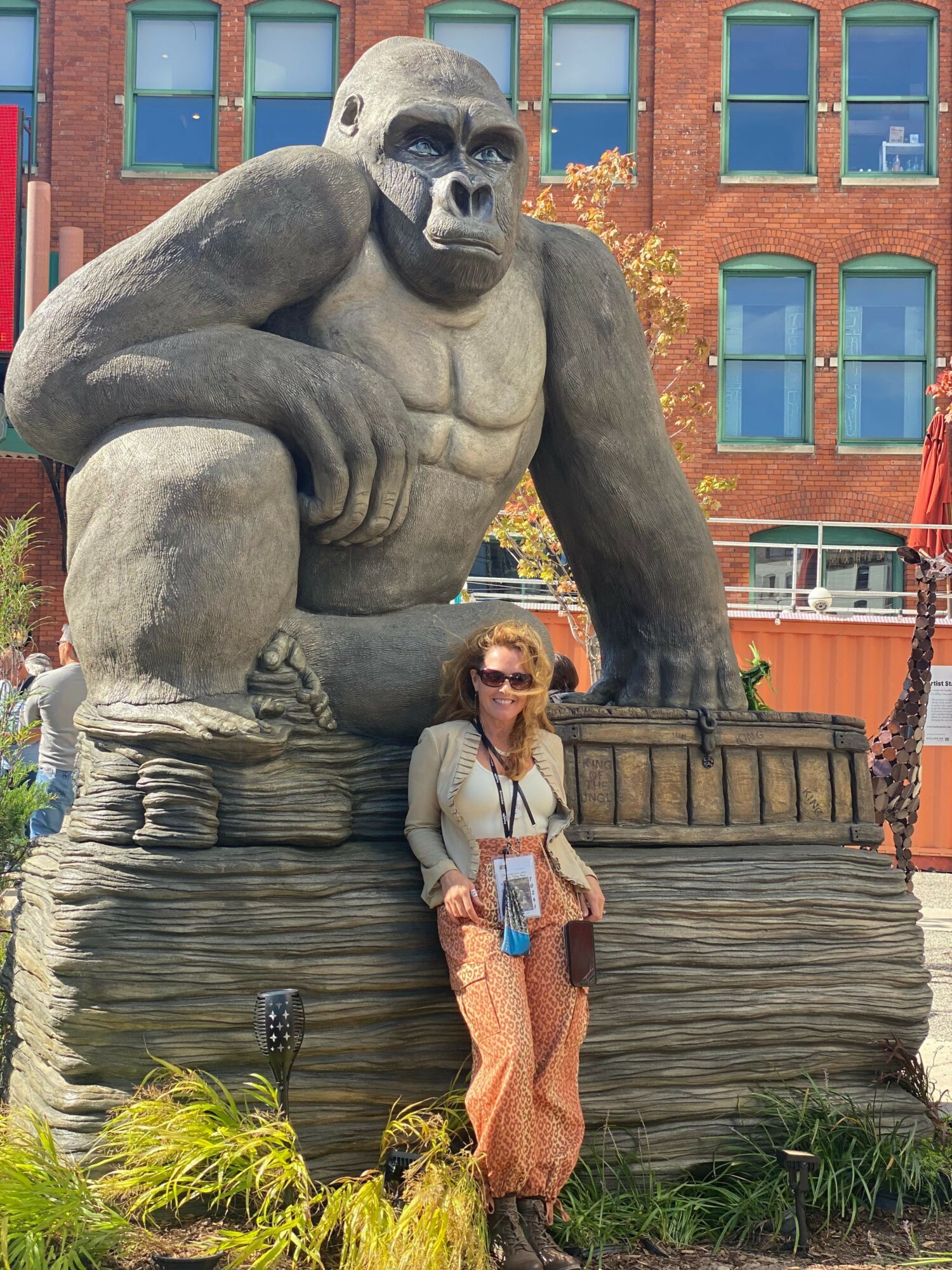 Person with long hair and sunglasses standing next to a large gorilla statue outdoors, with a brick building in the background.
