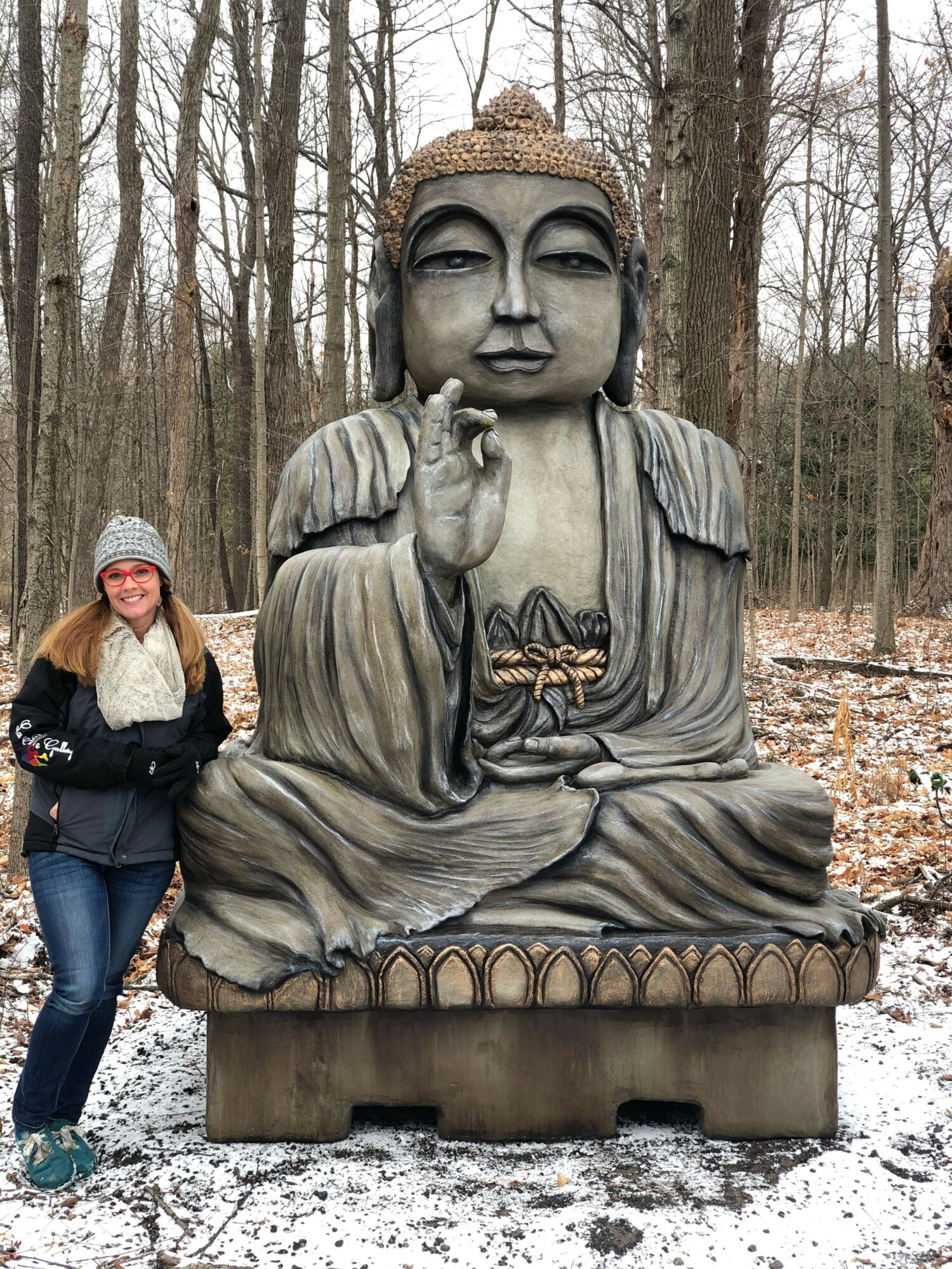 Person standing next to a large Buddha statue outdoors in a snowy forest, with trees in the background.