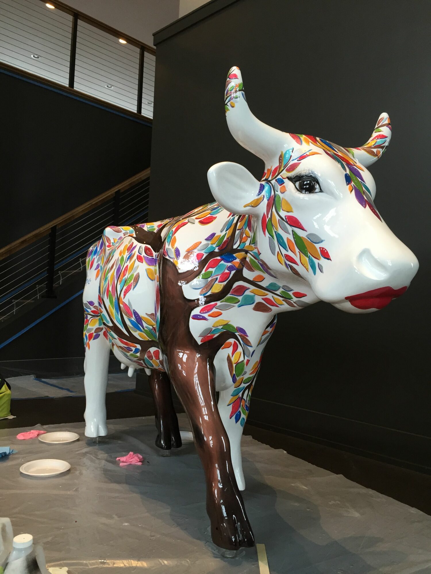 Colorful cow sculpture with a white body, multicolored patterns, and brown legs, standing on a gray floor.