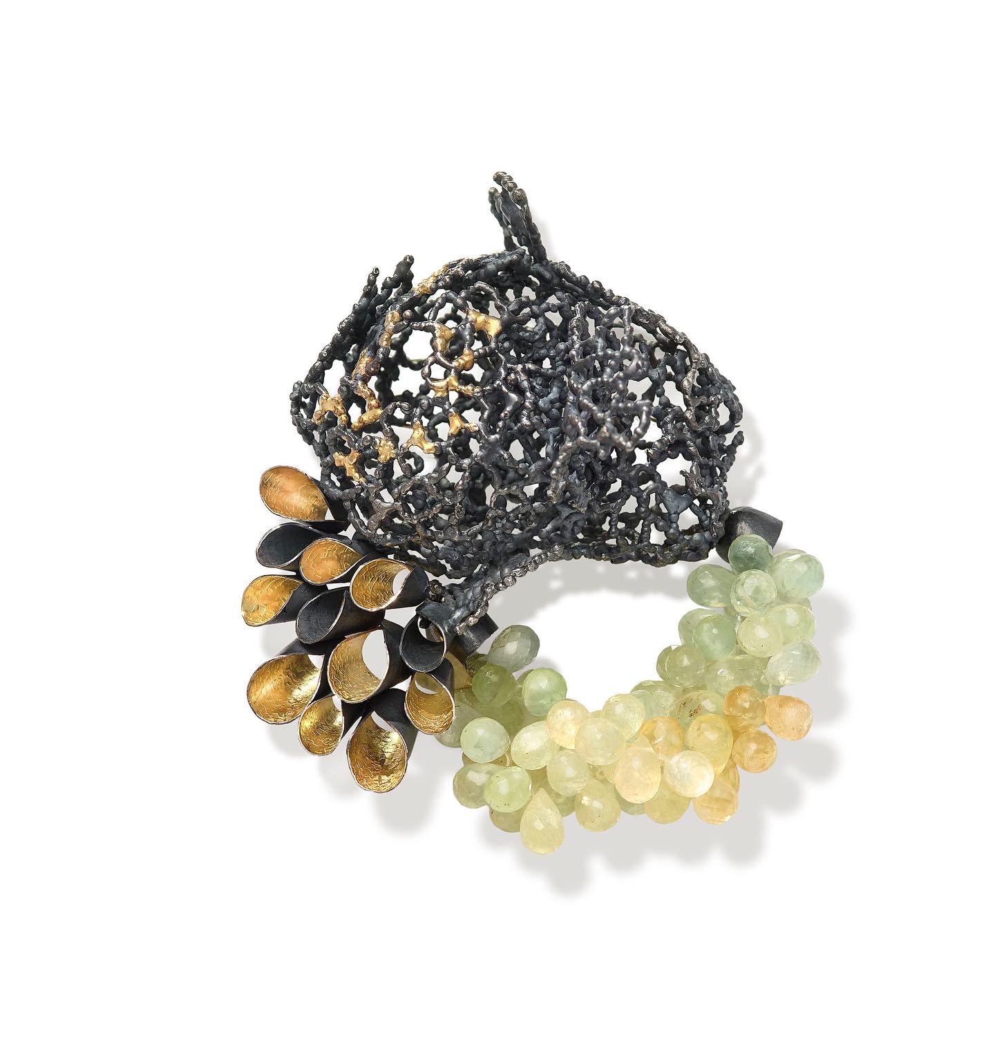 A black lace-like decorative object with beads and pearls, including gold, black, and light green beads, on a white background.