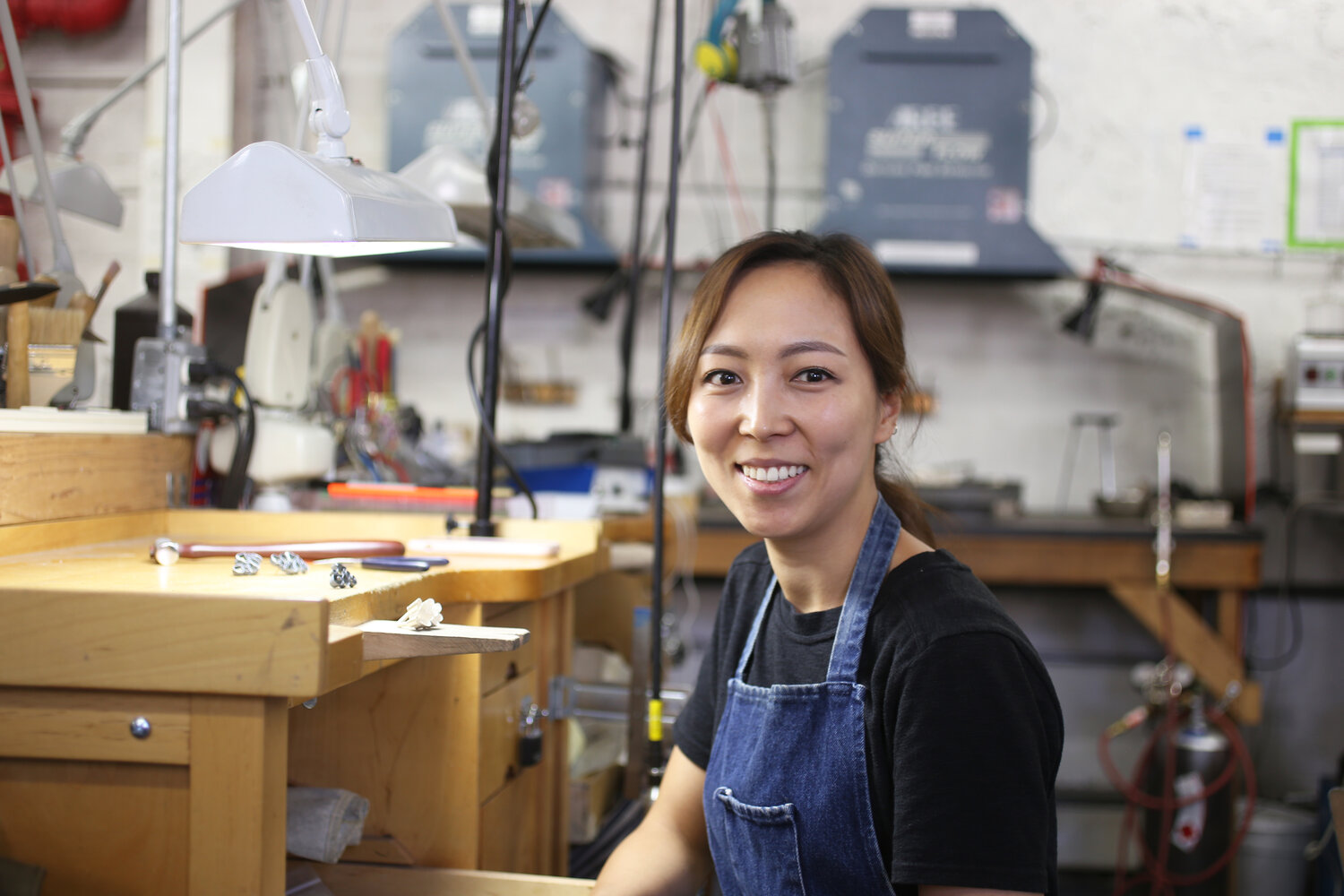 Young woman smiling in a workshop with tools and equipment, wearing a blue apron and black shirt.