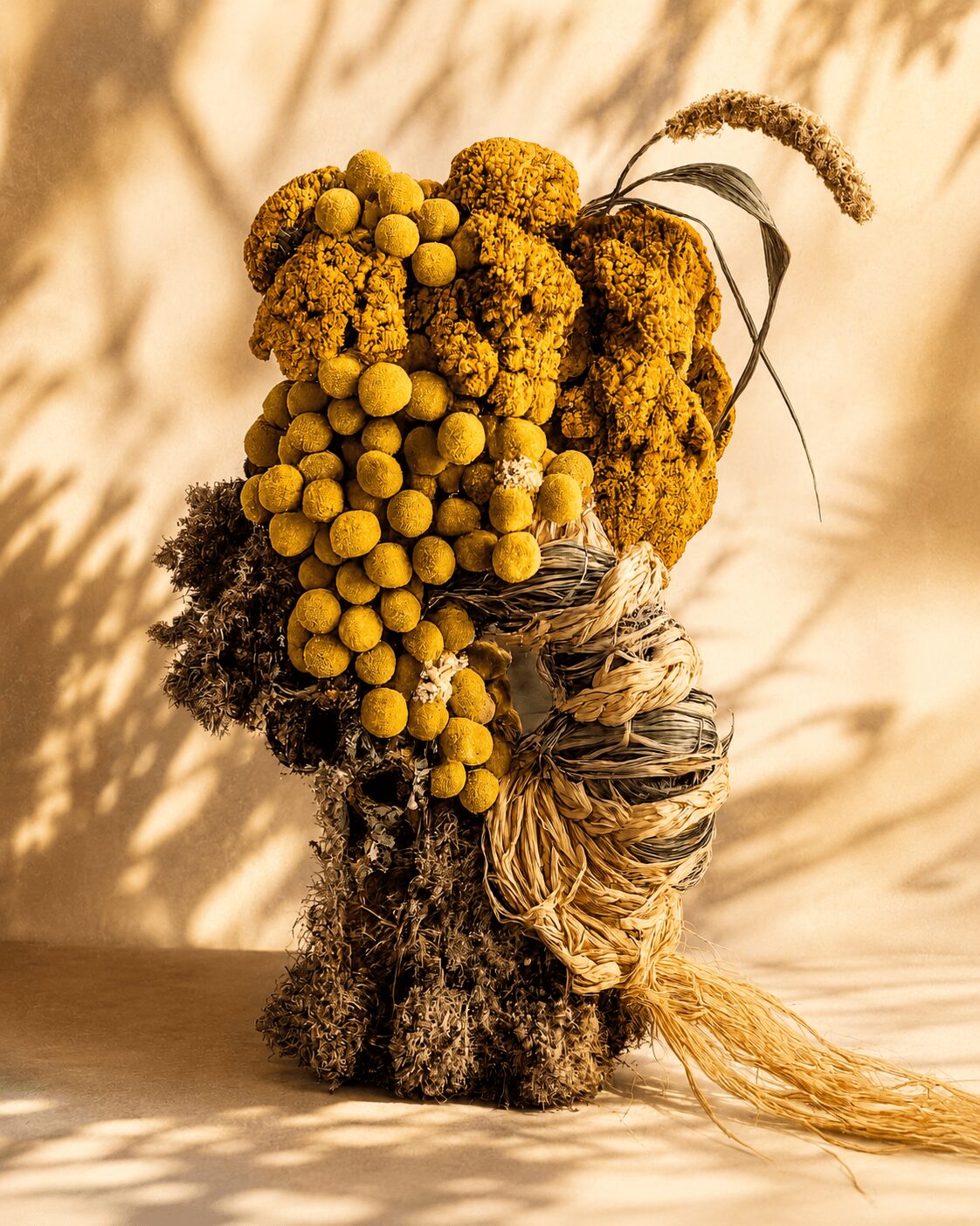 A bouquet of yellow and brown flowers and plants in a textured vase, with a light background and shadow pattern.