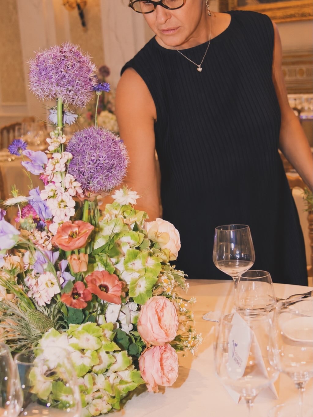 Woman in black dress arranging a large colorful flower bouquet on a table, with wine glasses and a water pitcher nearby.