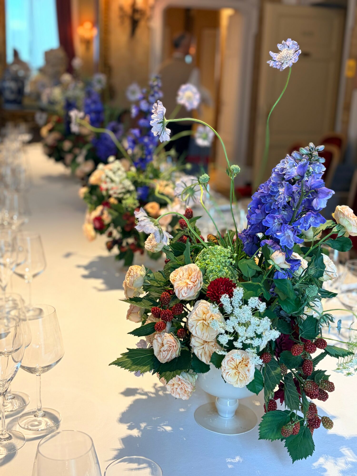 Elegant floral arrangement with purple, white, and pink flowers on a table, with glasses and a blurred background.