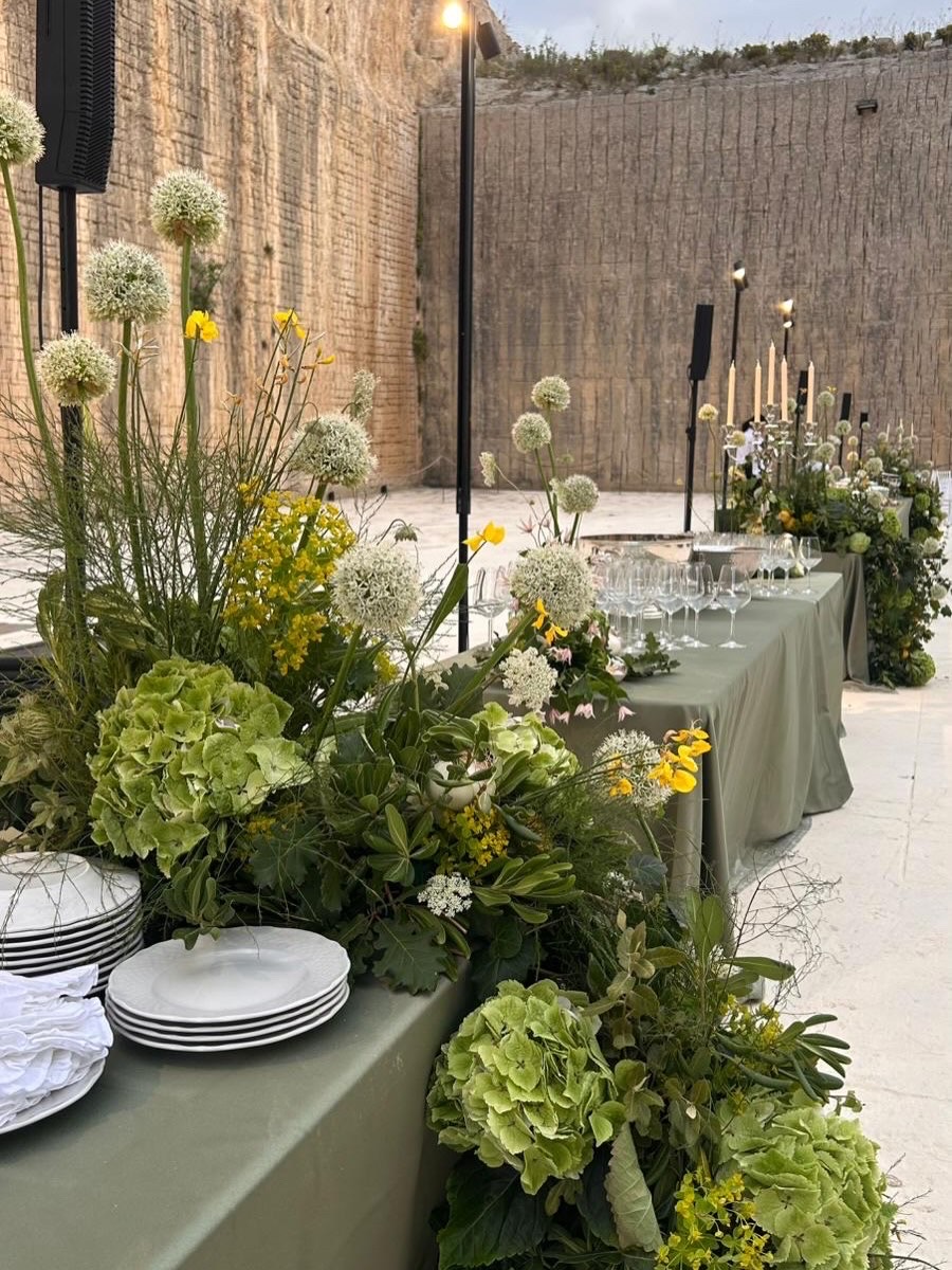 Outdoor table with floral arrangements, plates, and candles against a brick wall, set for an event.