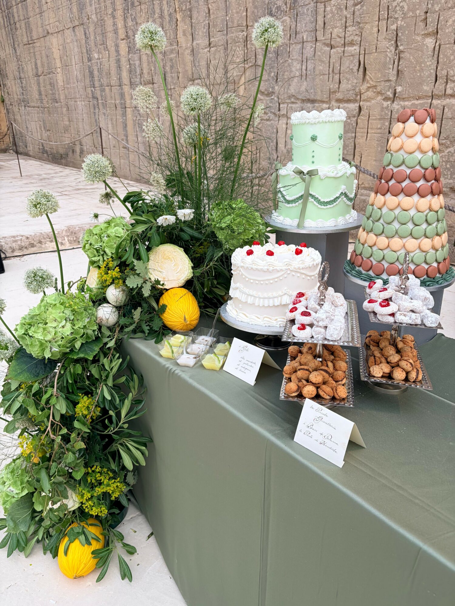 Table with a white cake decorated with red cherries, surrounded by flowers, nuts, and colorful desserts, against a brick wall.