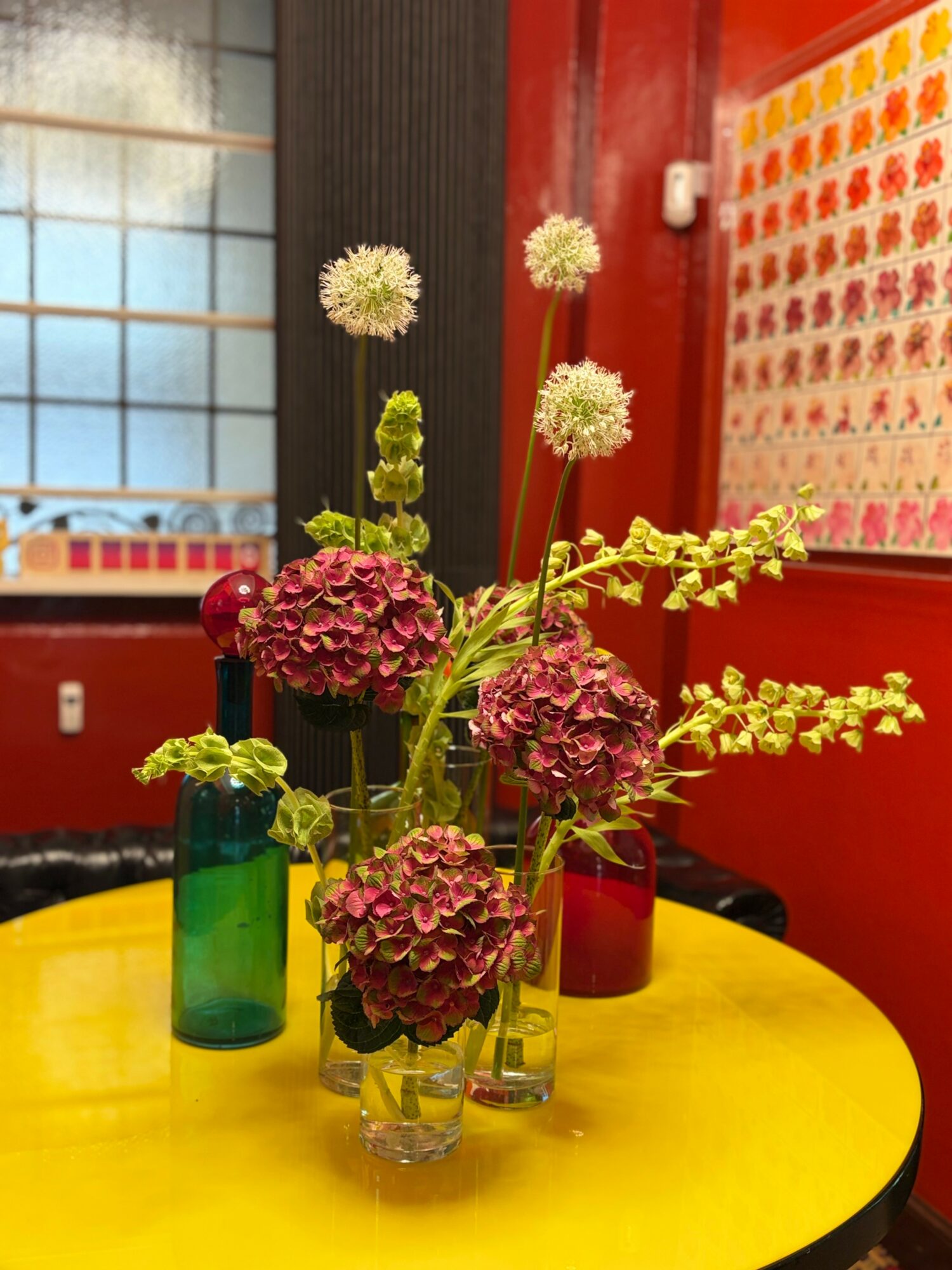 Vase with pink hydrangeas and tall white flowers on a yellow table, colorful wall in background.