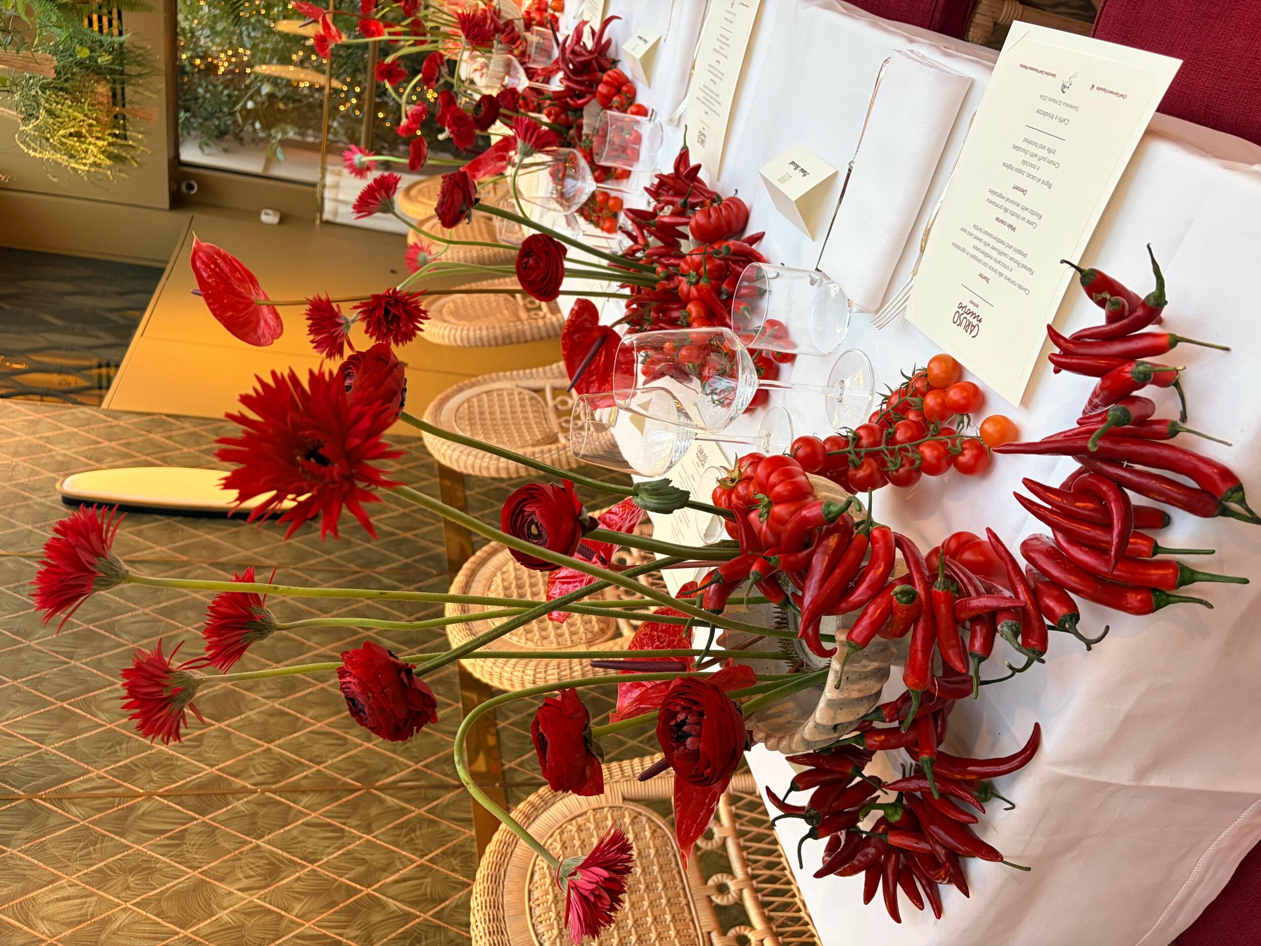 Table decorated with red flowers, chili peppers, and a menu, with chairs and a window in the background.