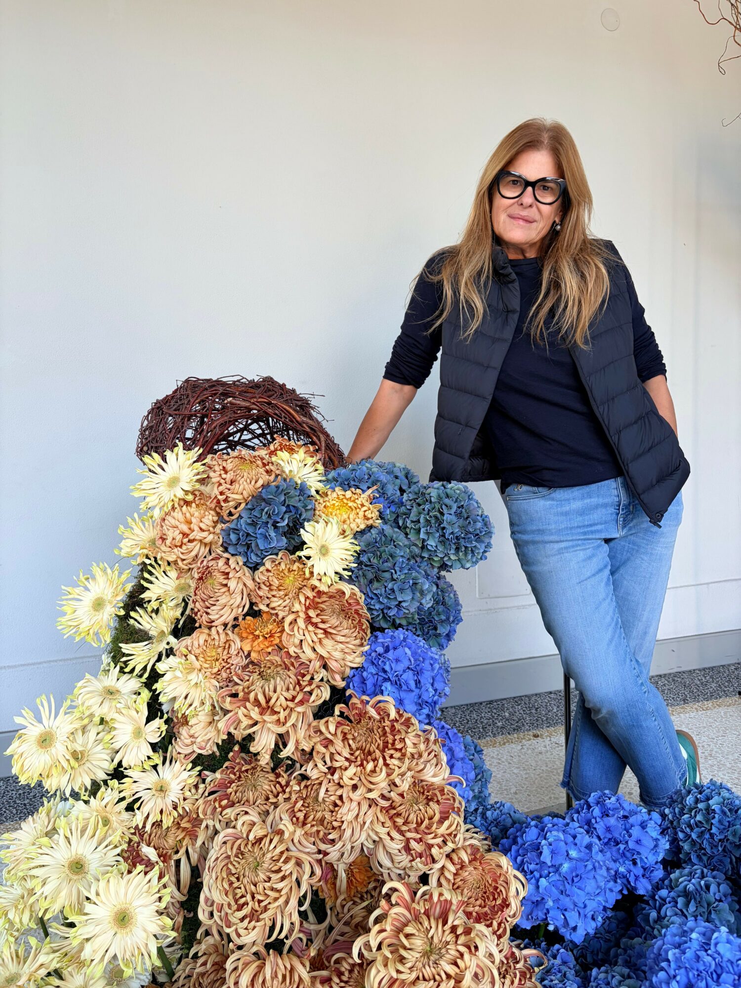 Woman with glasses and long hair standing next to a large arrangement of dried flowers, wearing a black jacket and jeans.