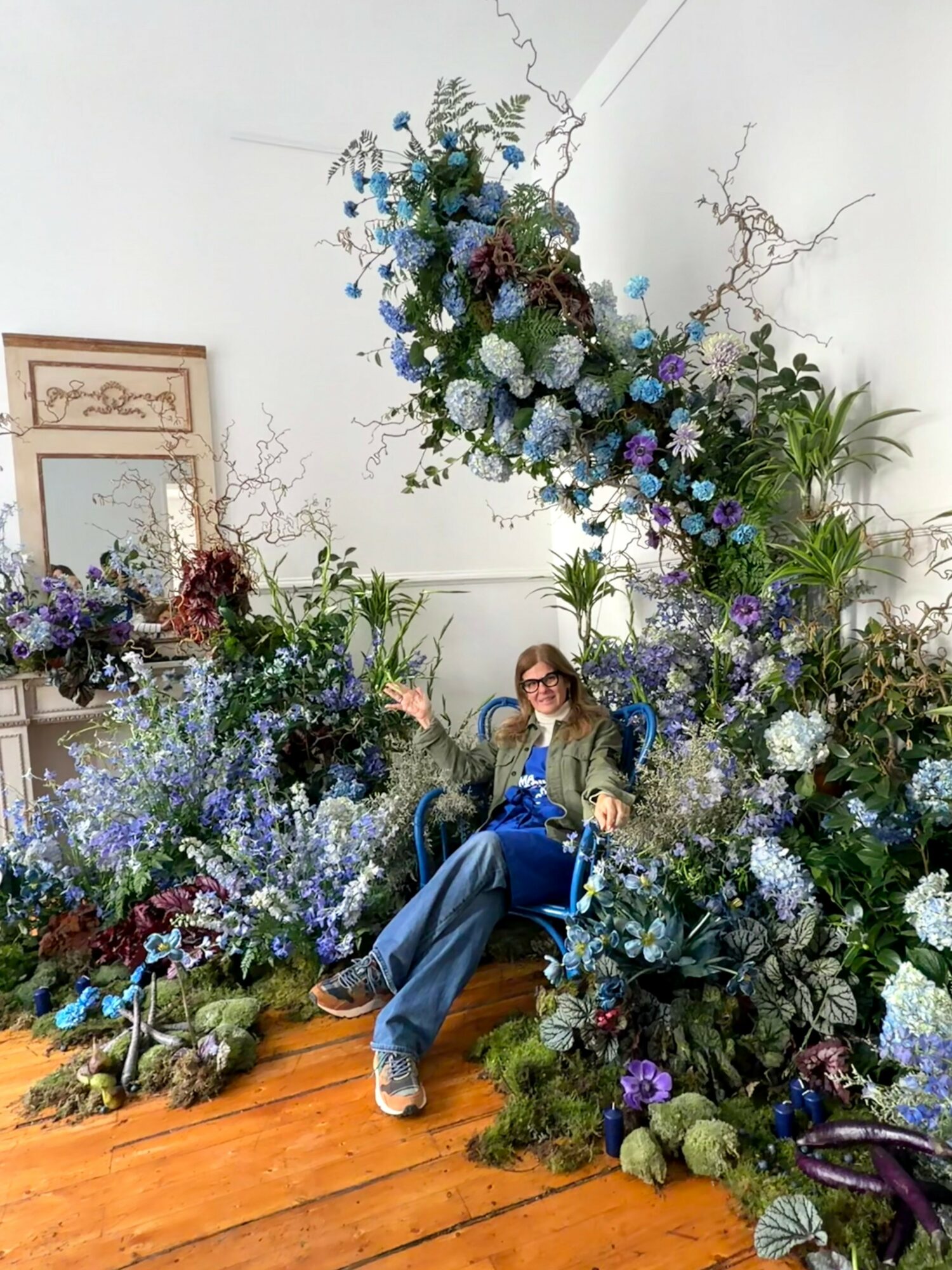Person sitting among large blue and purple floral arrangements with greenery, in an indoor space with a mirror and white wall.
