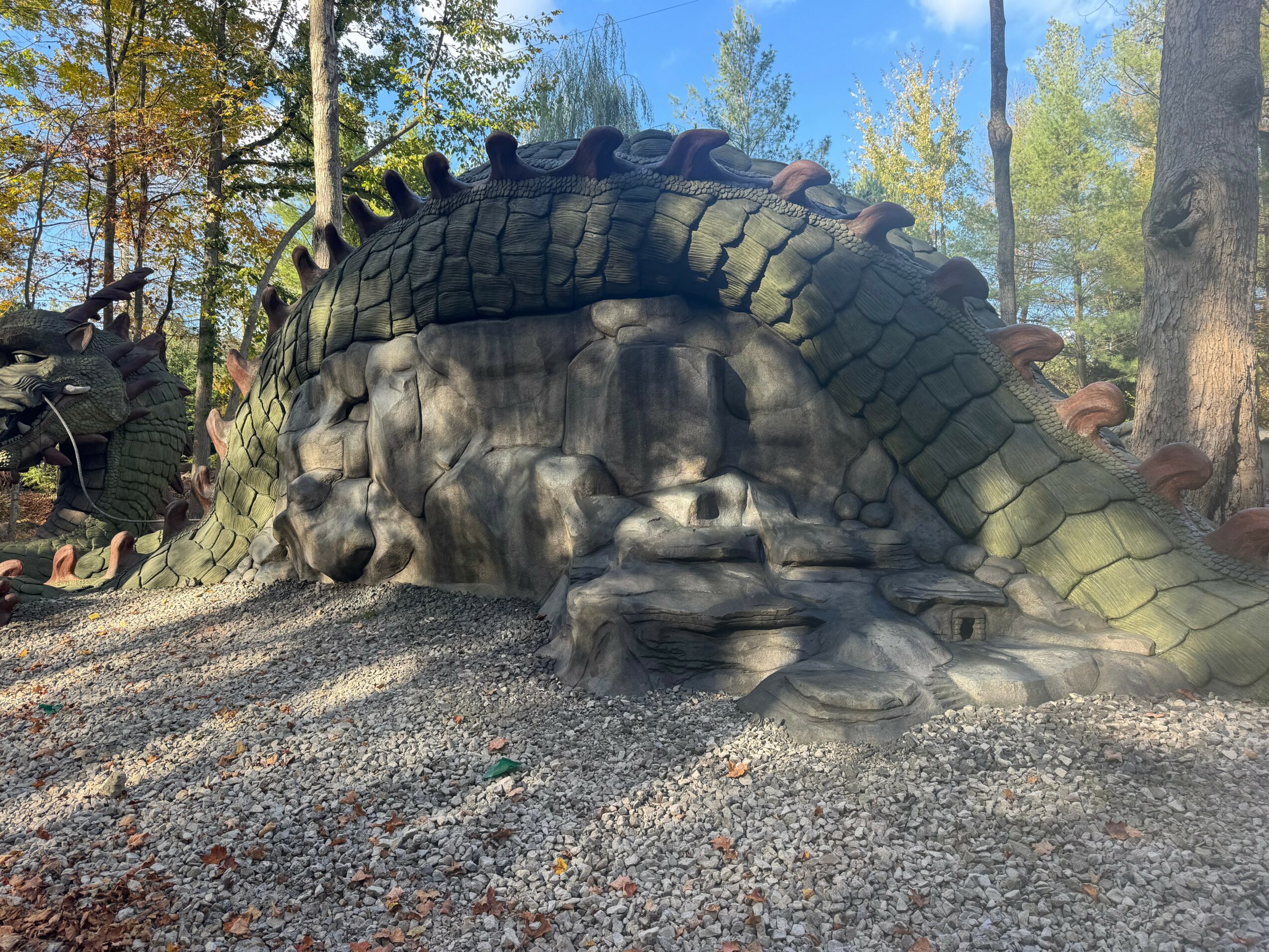 Dinosaur sculpture outdoors with trees and blue sky in background, resting on gravel ground.