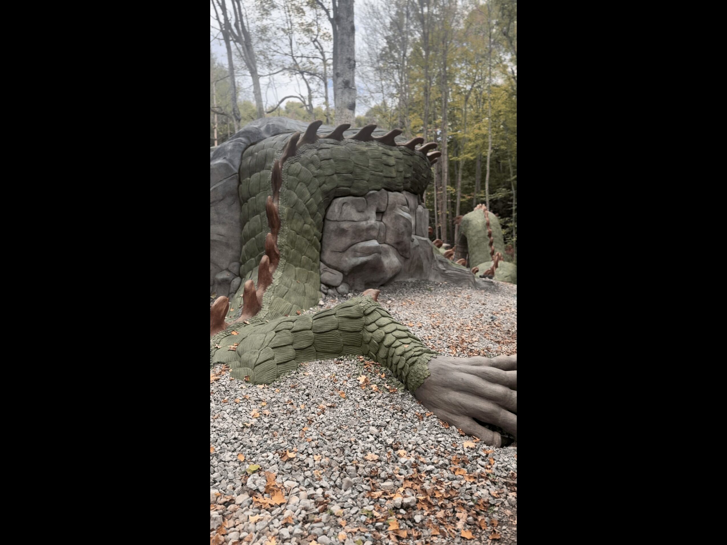 A large crocodile sculpture with an open mouth, surrounded by trees and a person in the background, on a gravel surface.