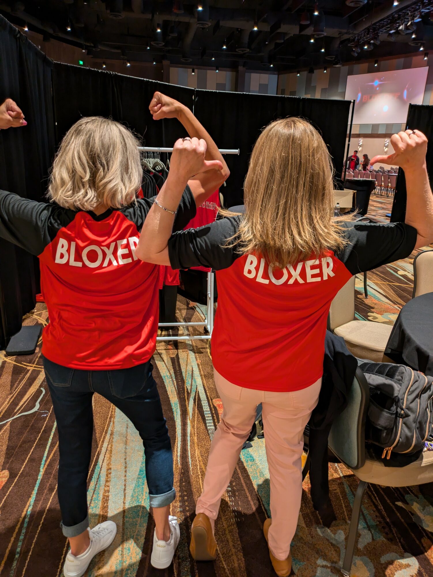Two children with blonde hair wearing red shirts with 'BLOXER' text, flexing their arms in a conference room.