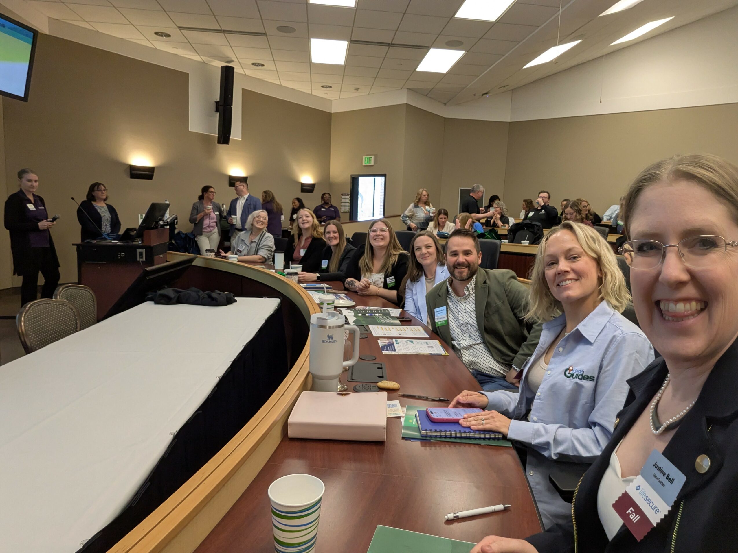 Group of people sitting at a conference table, smiling, with others standing in the background, in a large room.