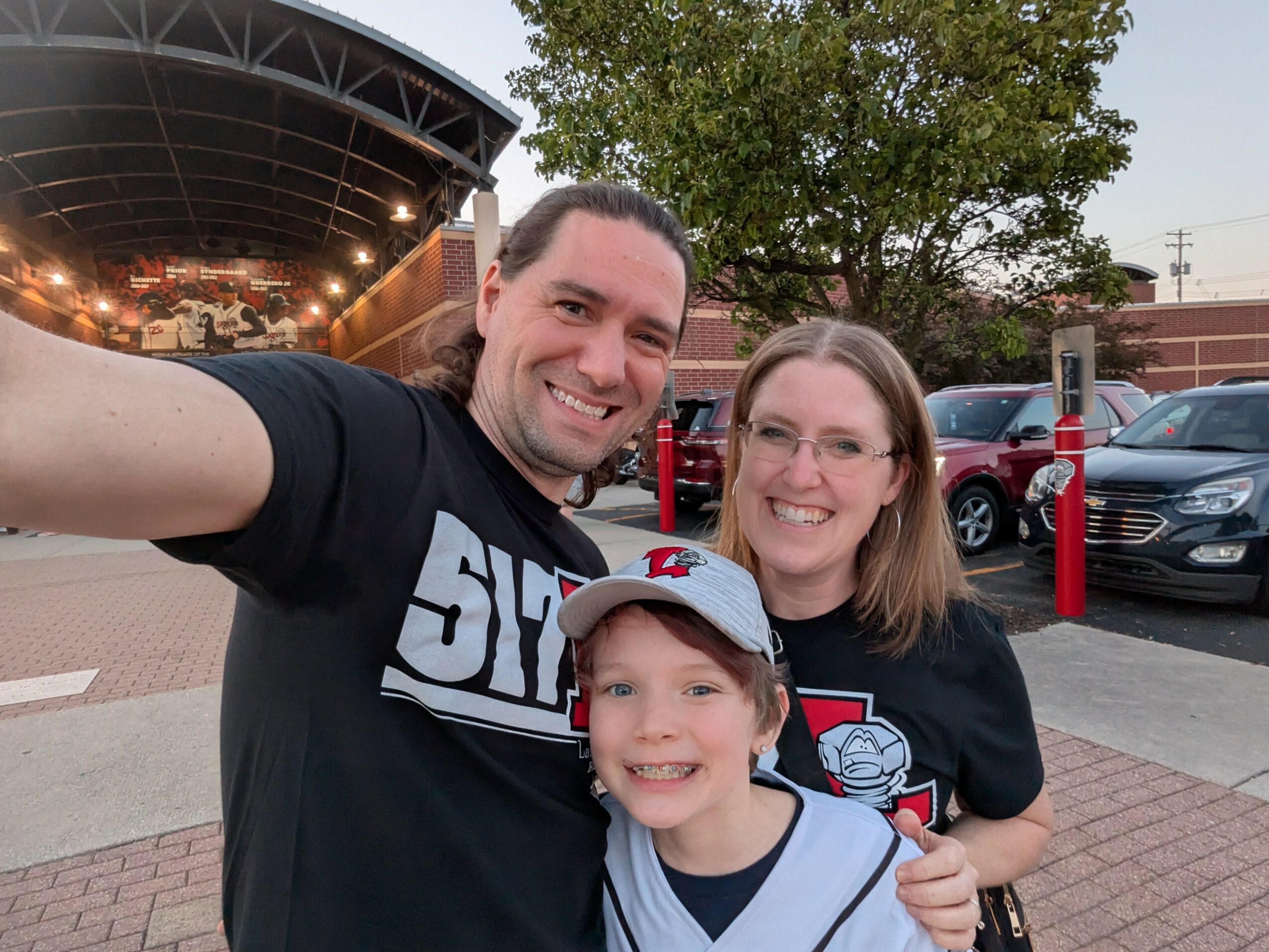 A smiling man, woman, and child pose outdoors, with trees and parked cars in the background.