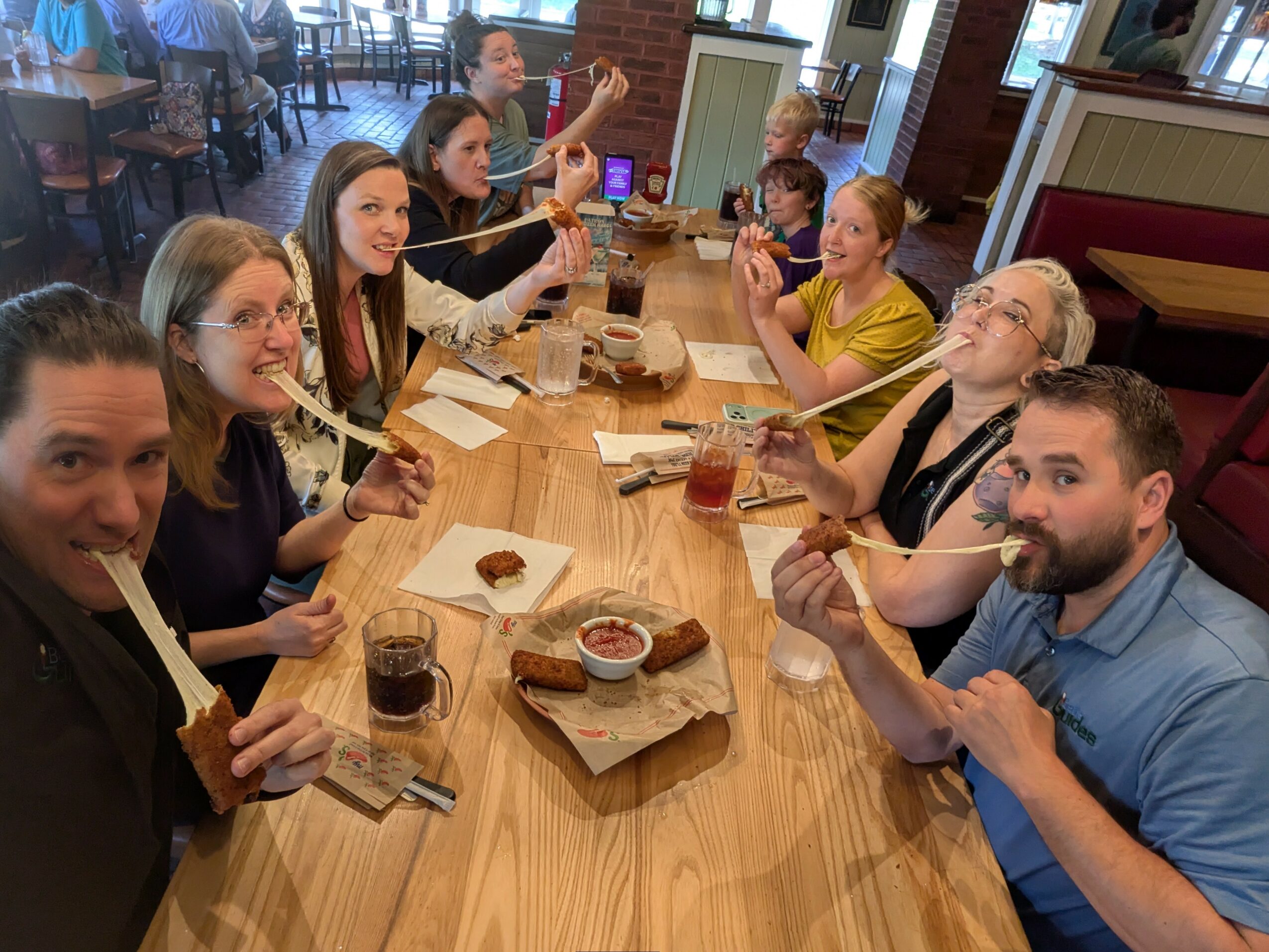 Group of people sitting at a long table in a restaurant, eating and drinking, smiling and engaging with each other.
