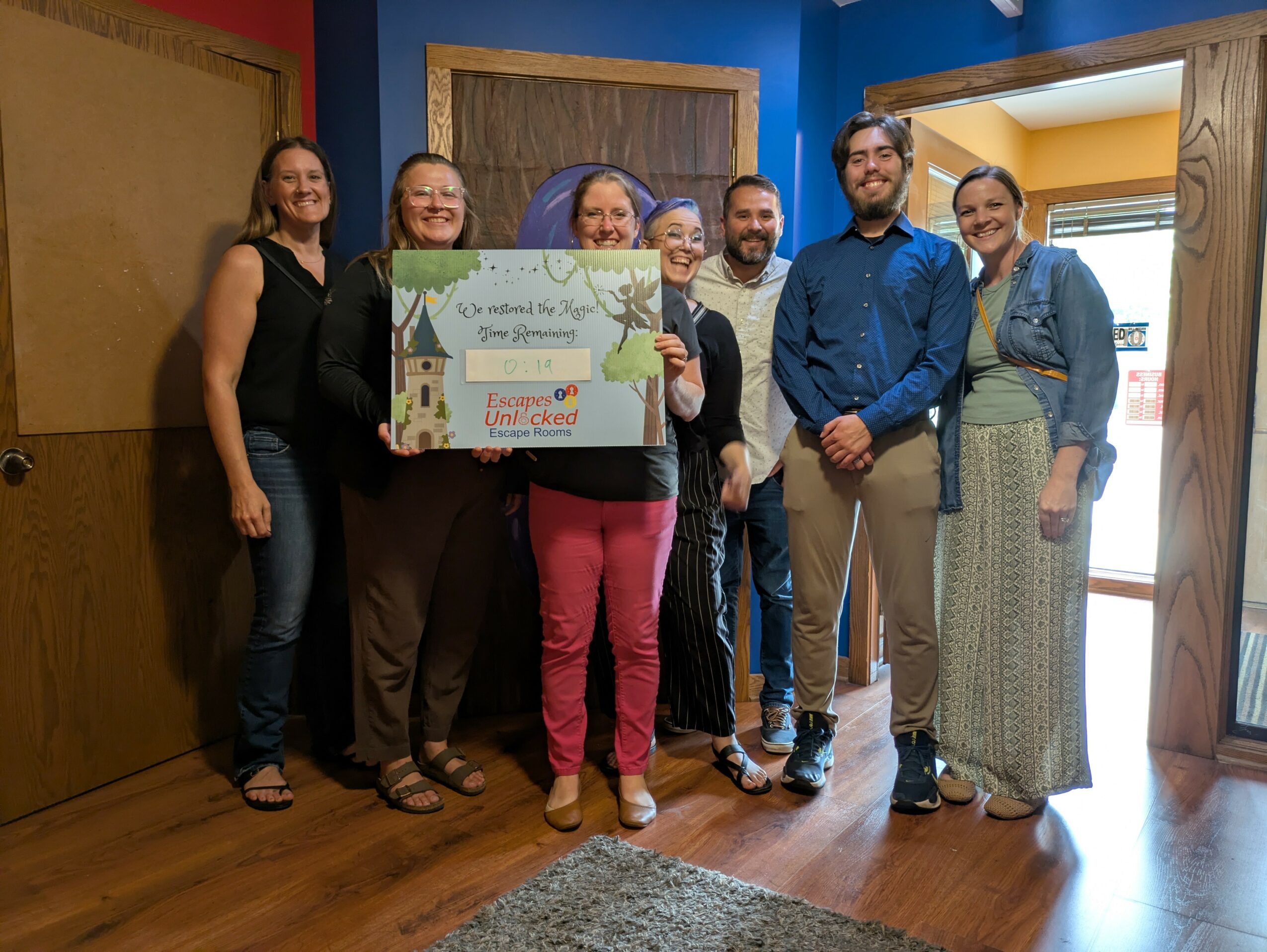 Group of seven people standing indoors, holding a large check, smiling, with wooden walls and a window in the background.