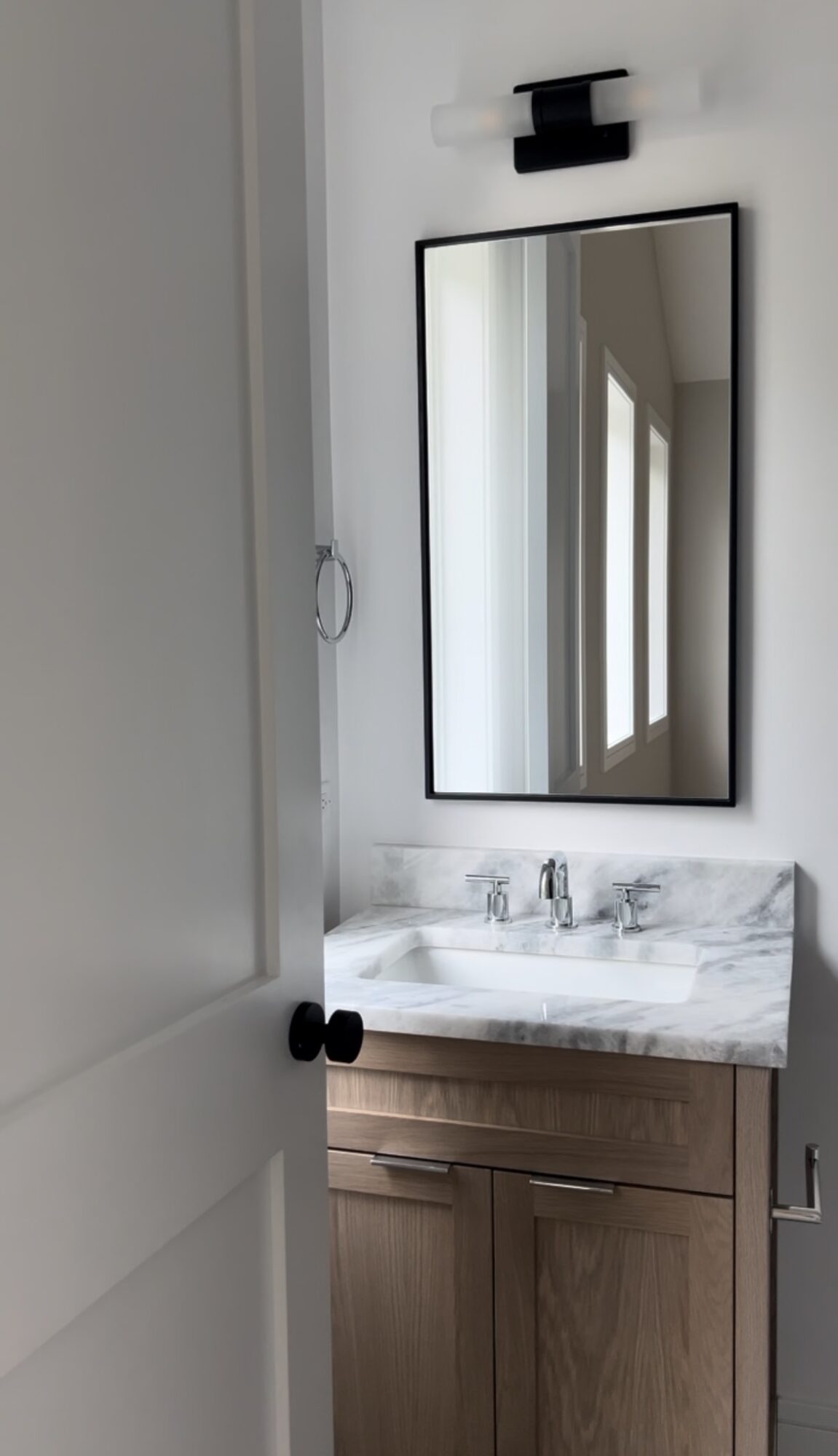 Bathroom mirror above sink with marble countertop and wooden cabinet, reflected in the mirror, with a wall-mounted light fixture above.