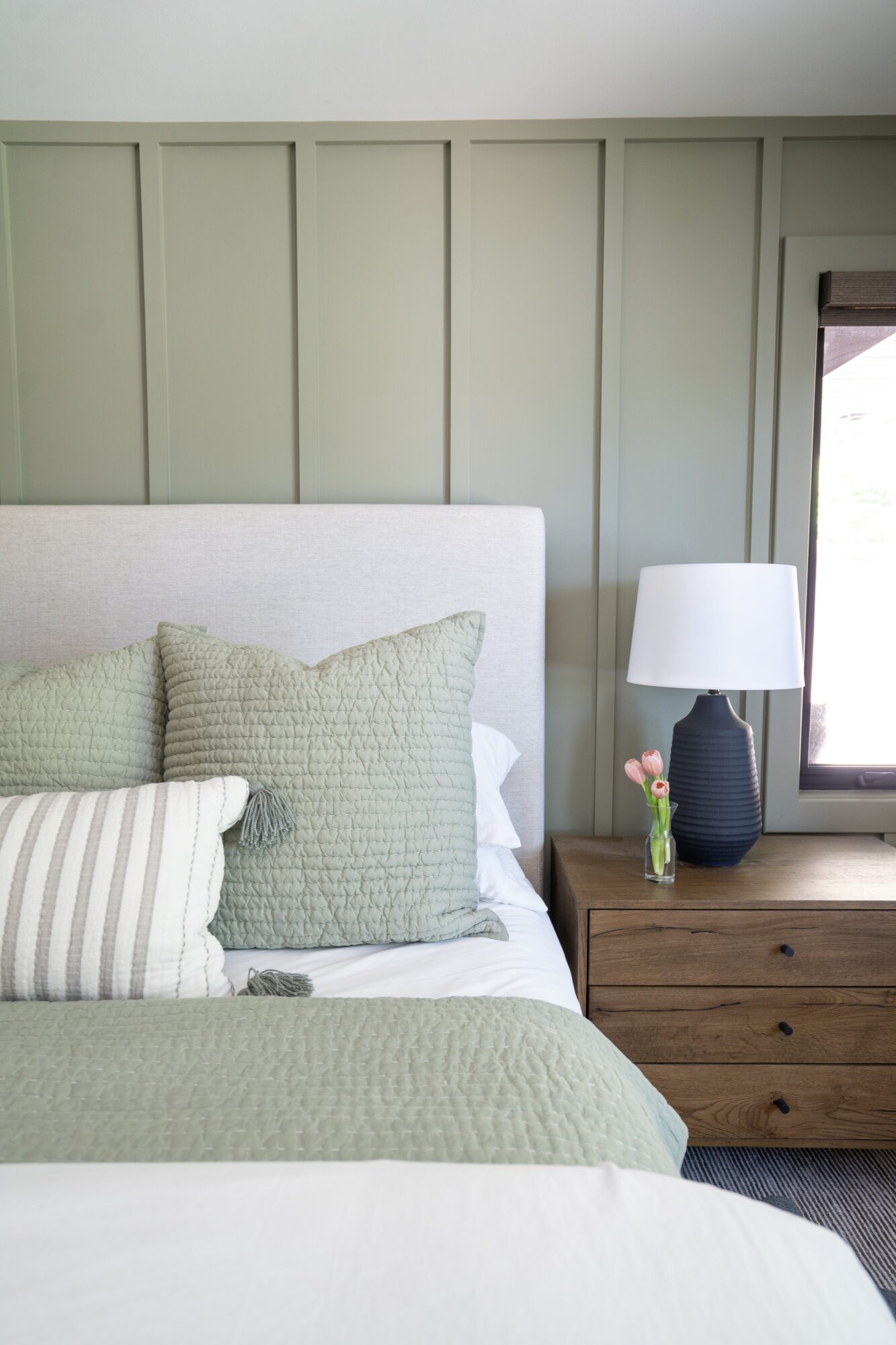 Bedroom with bed, pillows, nightstand, and lamp, against a paneled wall and window, in neutral tones.
