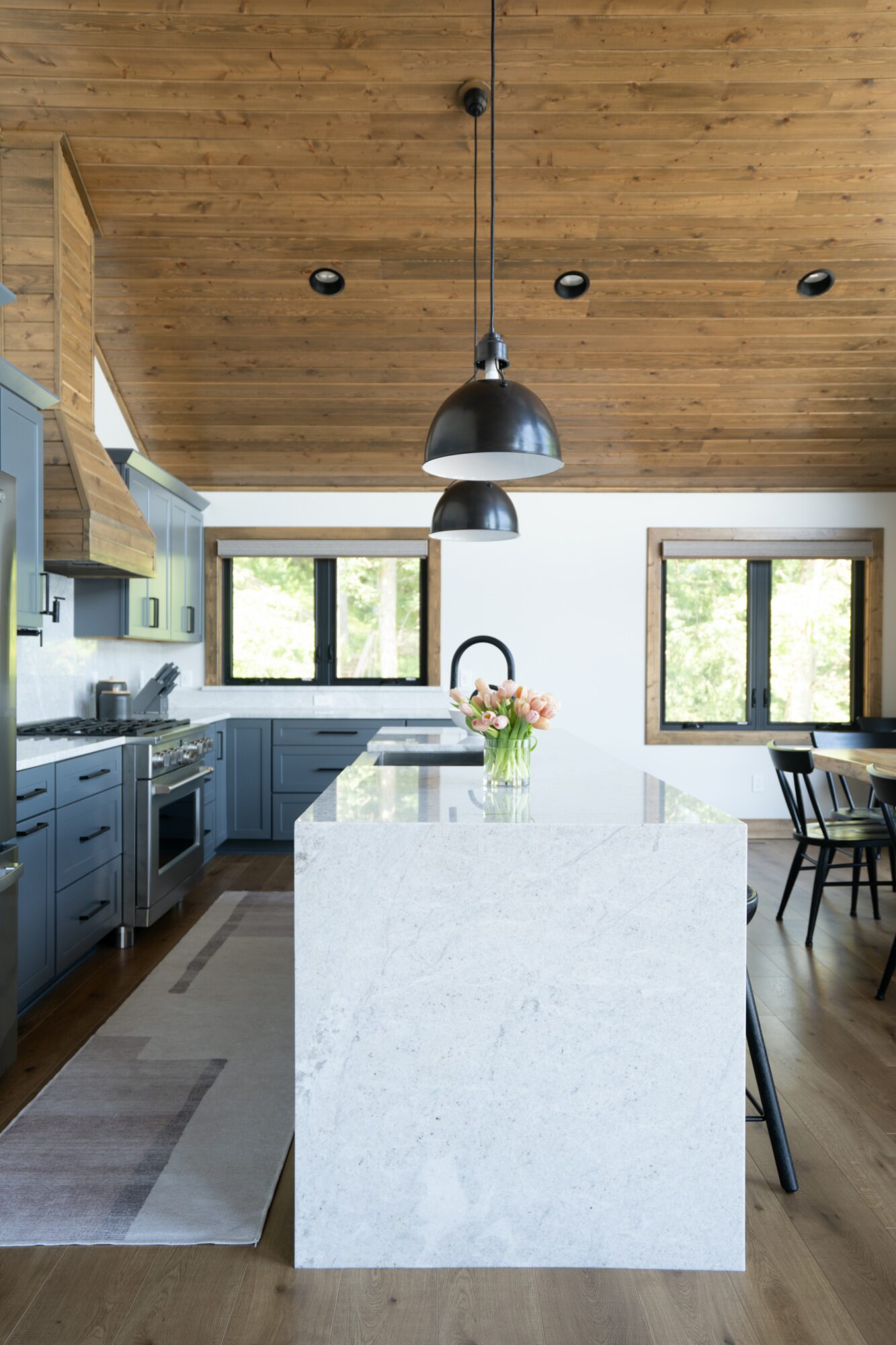Modern kitchen with wooden ceiling, white island, black pendant light, and large windows, with dining area nearby.