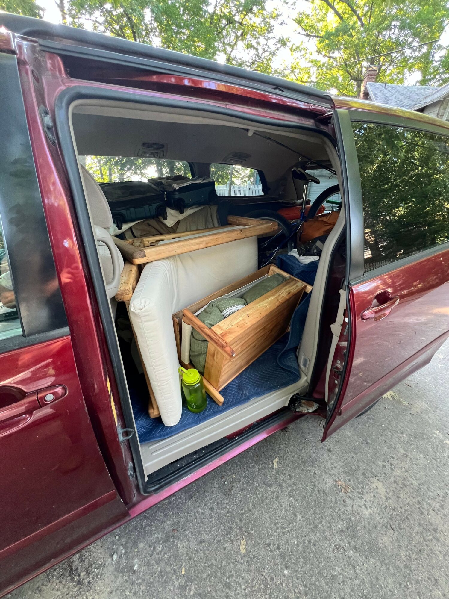 Red car with open trunk, containing a wooden chair and a small table inside, parked on a street with trees nearby.
