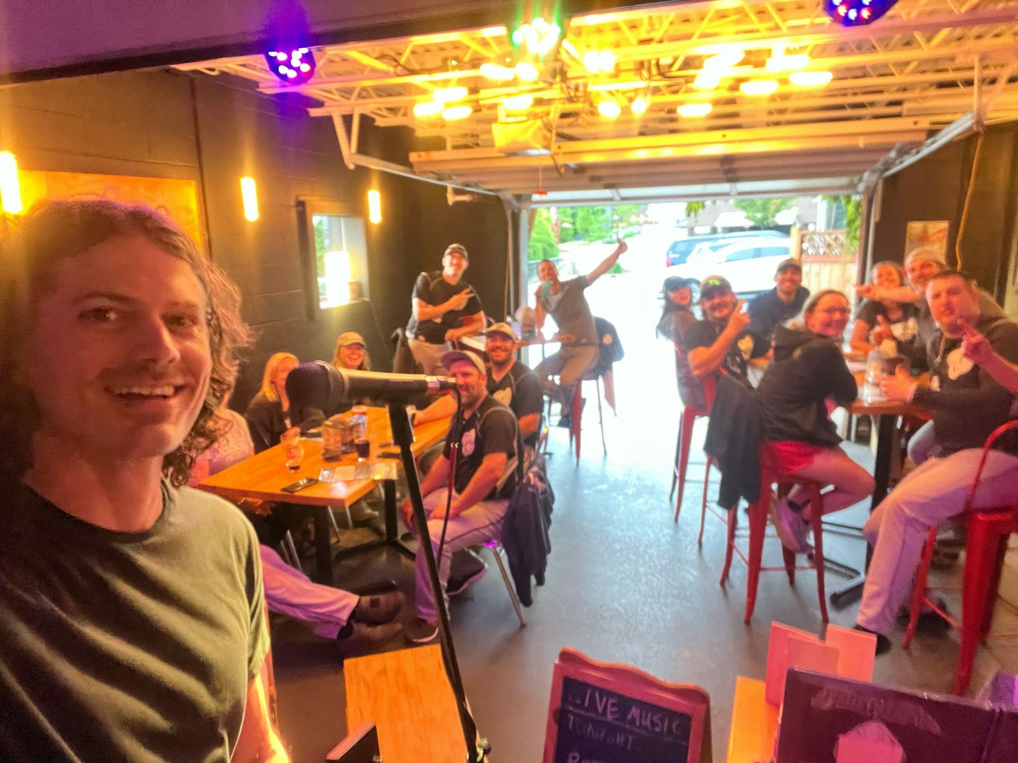 People sitting at tables in a restaurant or cafe, with a woman smiling at the camera, warm lighting, and a lively atmosphere.