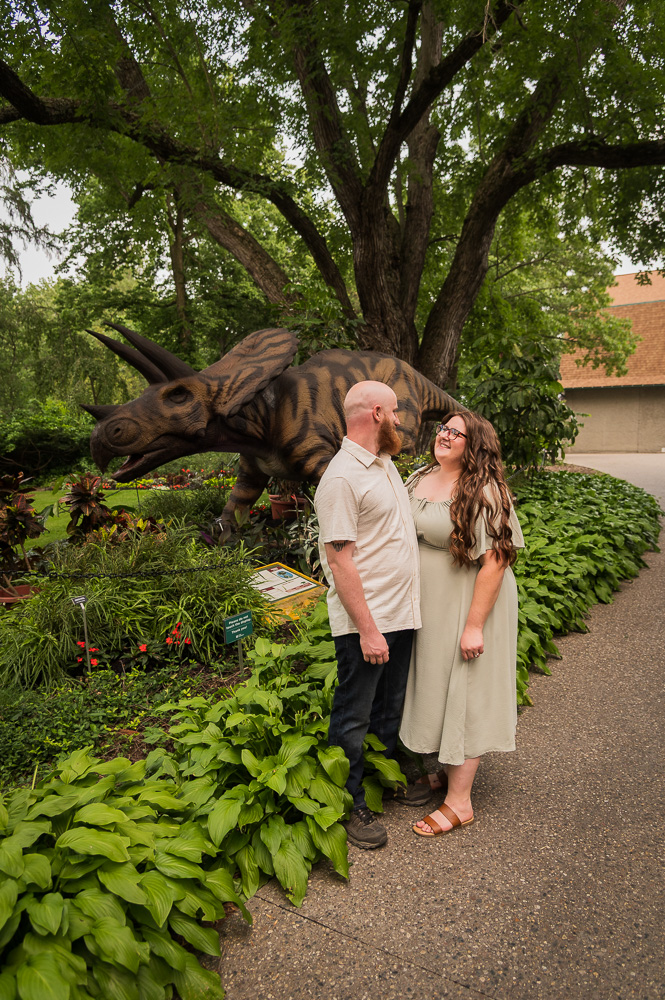Two people stand near a dinosaur sculpture outdoors, surrounded by greenery and a large tree, with a building in the background.