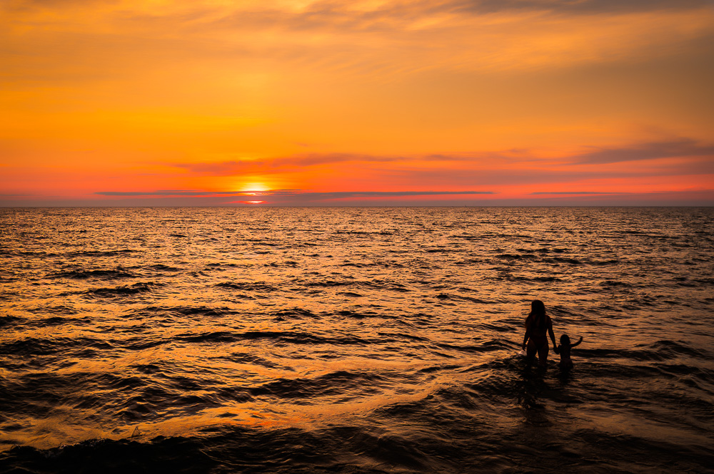 Sunset over the ocean with two people standing in shallow water, silhouetted against the colorful sky.