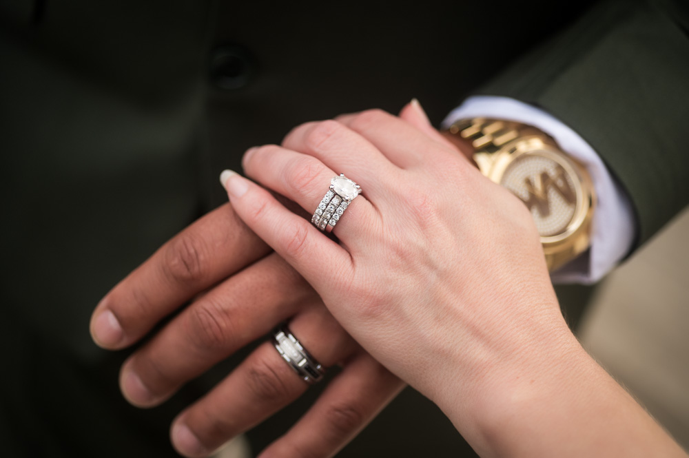 Two hands with wedding rings, one hand resting on the other, with a blurred background, one person wearing a watch.