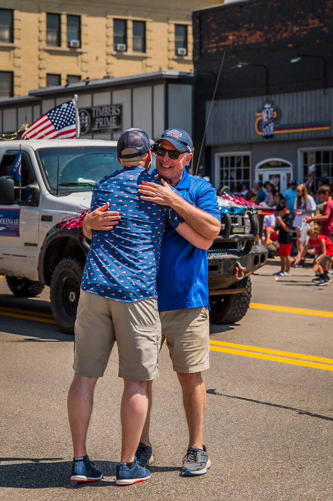 Two men hugging on a street with people, vehicles, and buildings in the background.