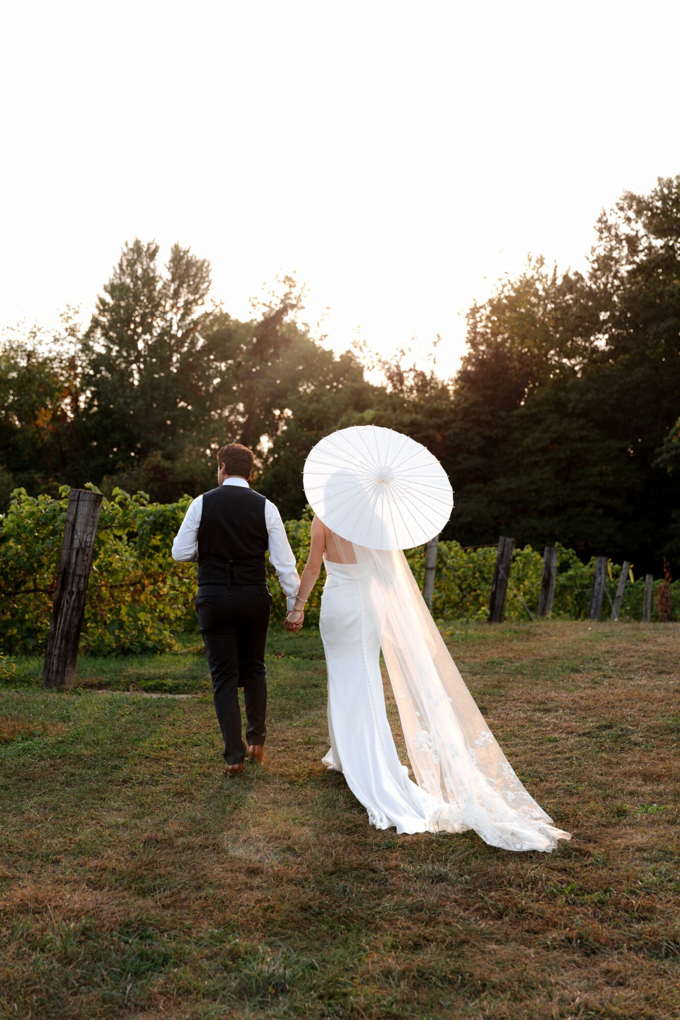 A couple walking outdoors, the woman holding a white parasol, surrounded by trees and greenery, with a bright sky above.