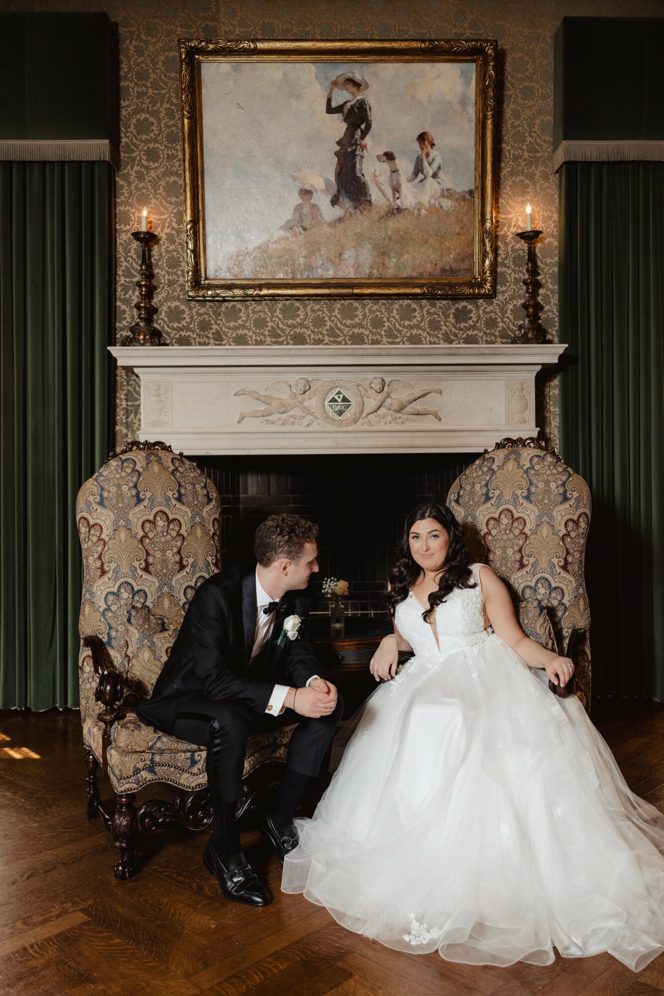 A couple in formal attire sitting on ornate chairs in front of a fireplace with a painting above, in a decorated room.
