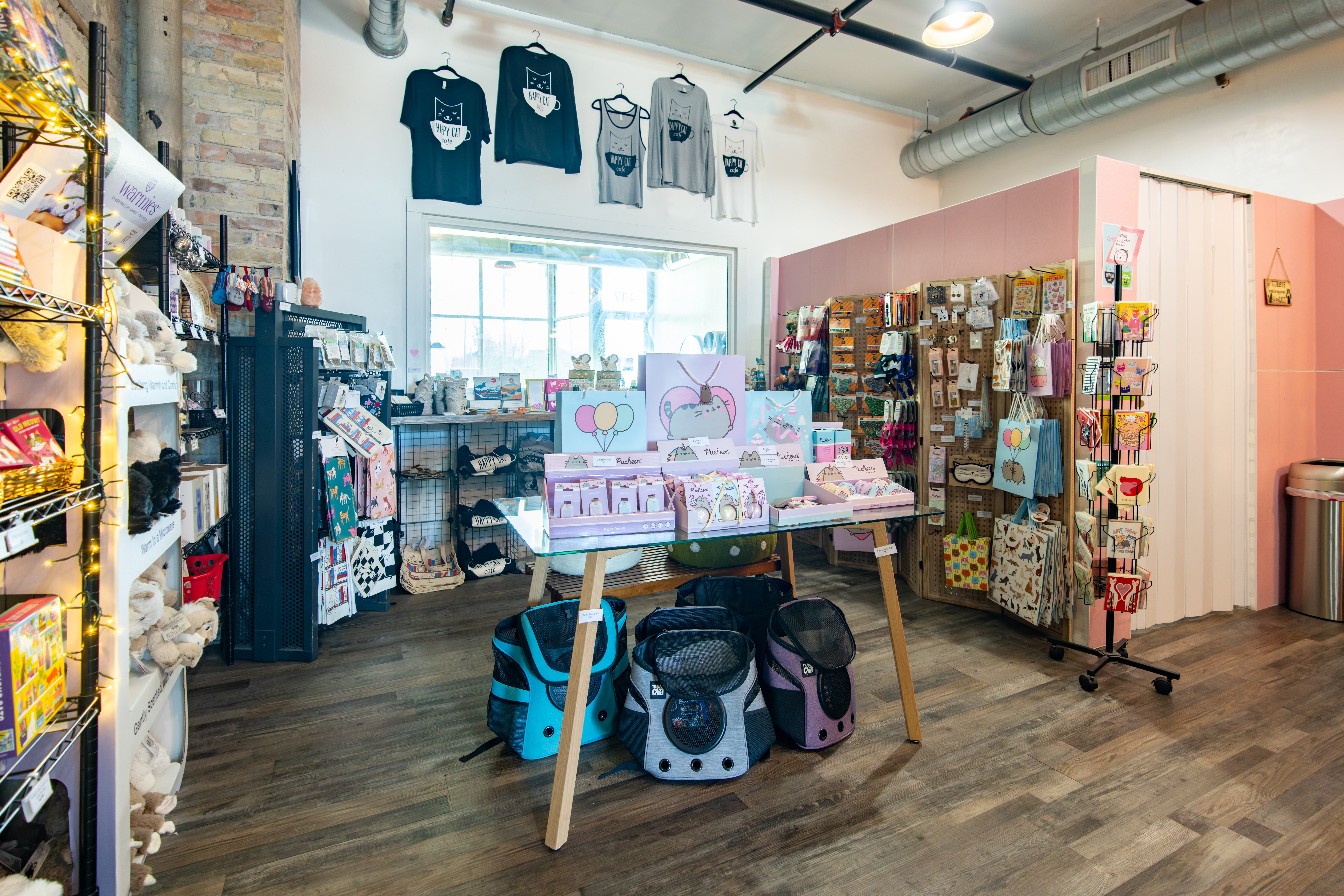 Display of clothing, accessories, and merchandise in a retail store with shelves and a table, wooden flooring, and a window.