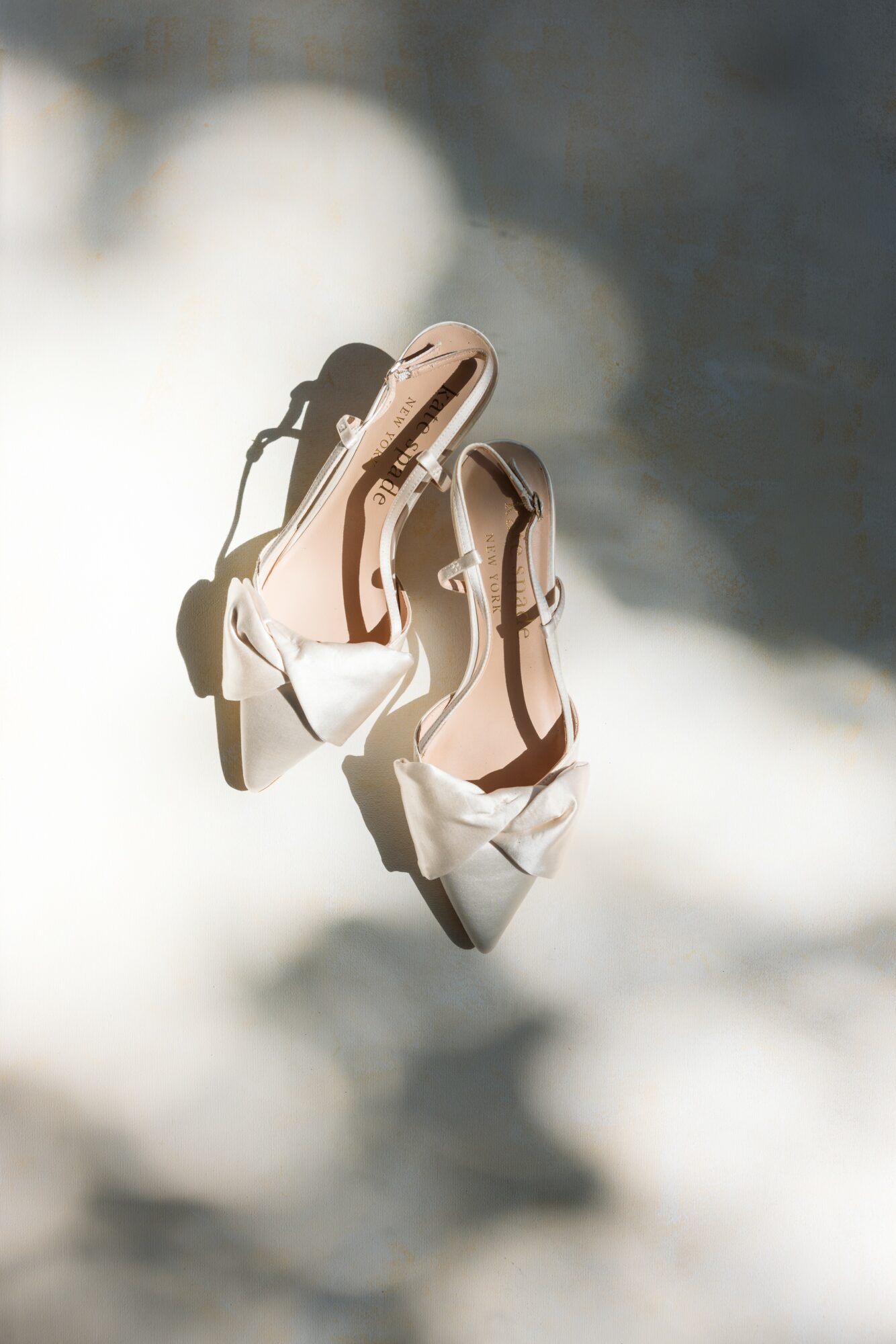 Pair of white high-heeled shoes with bows, placed on a surface with shadows and light spots