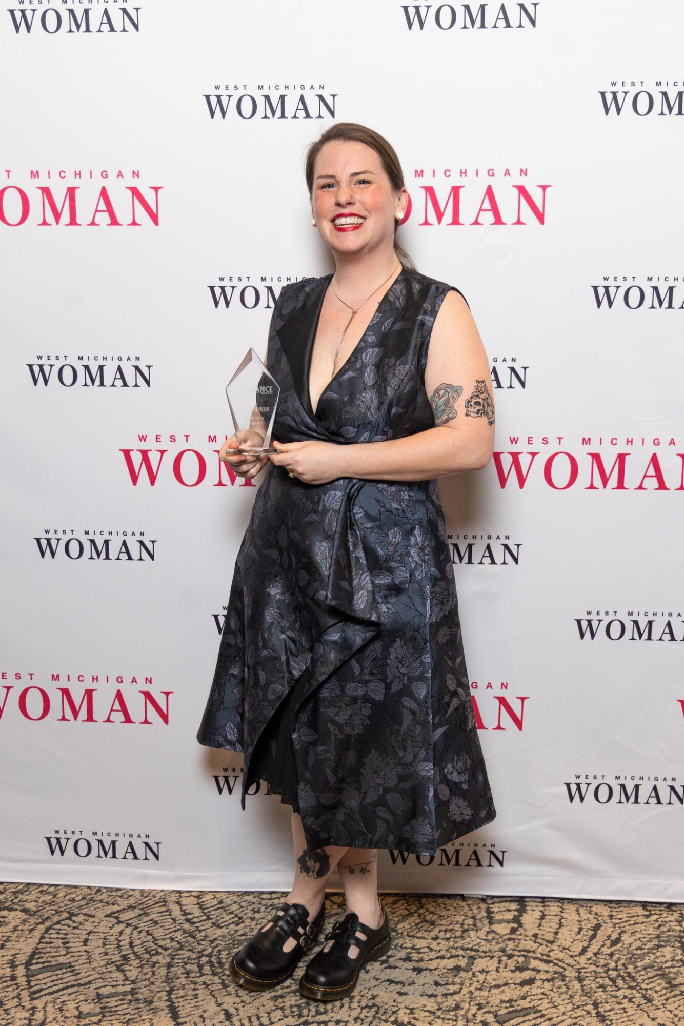 Woman in a dark floral dress holding an award, smiling, standing in front of a backdrop with text, wearing black shoes.