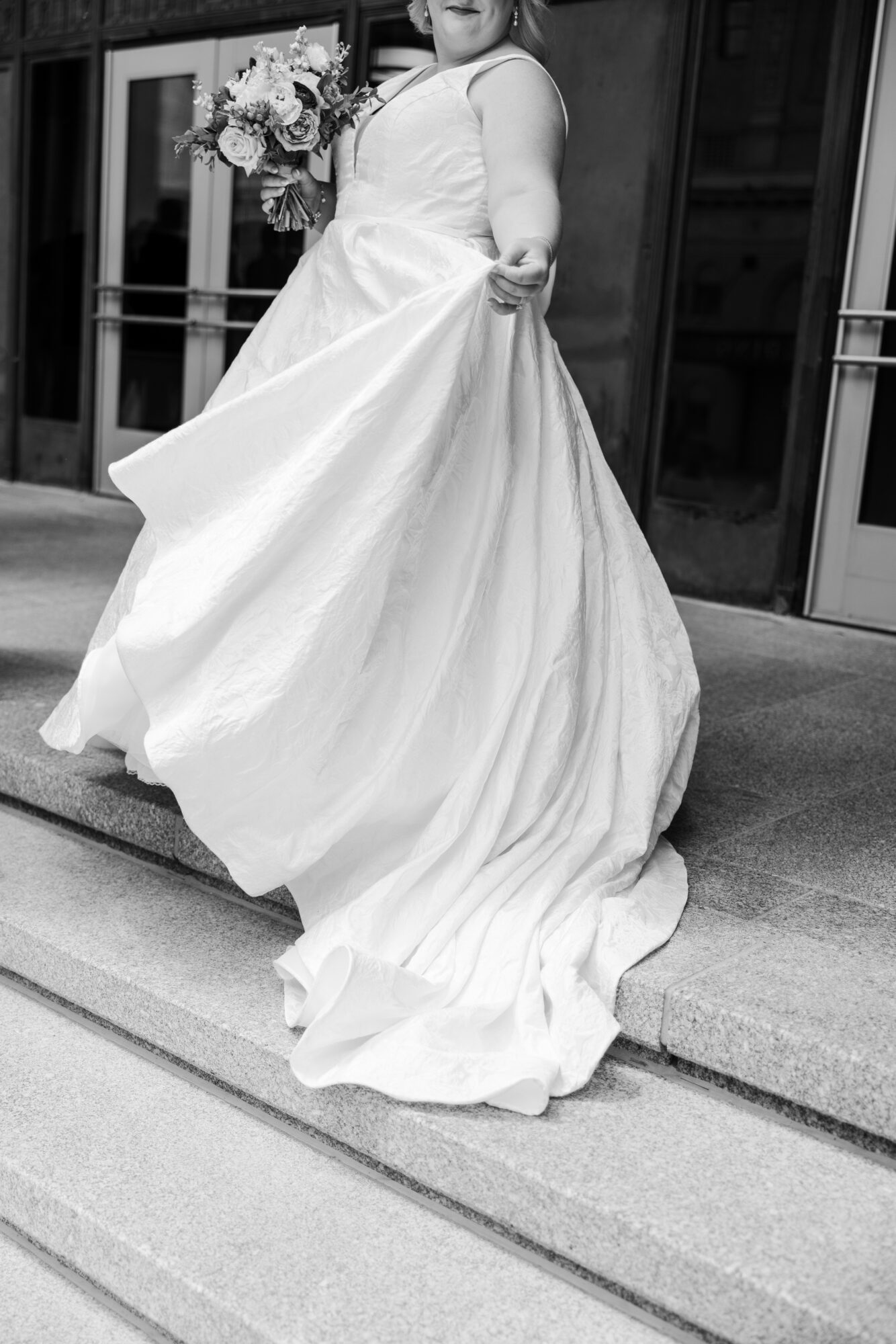 Person in wedding dress holding a bouquet, standing on steps outside building, black and white photo.