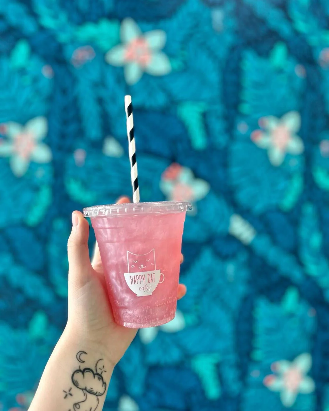 Hand holding a pink drink in a clear cup with a striped straw, background of blue floral pattern.