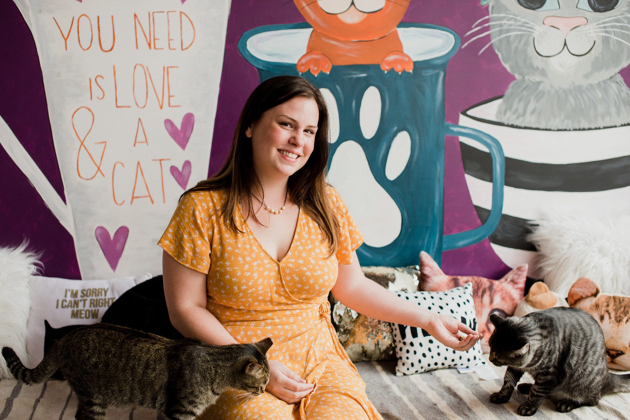 Smiling woman in yellow dress sitting on a couch with two cats, colorful mural background with cartoon animals and text.
