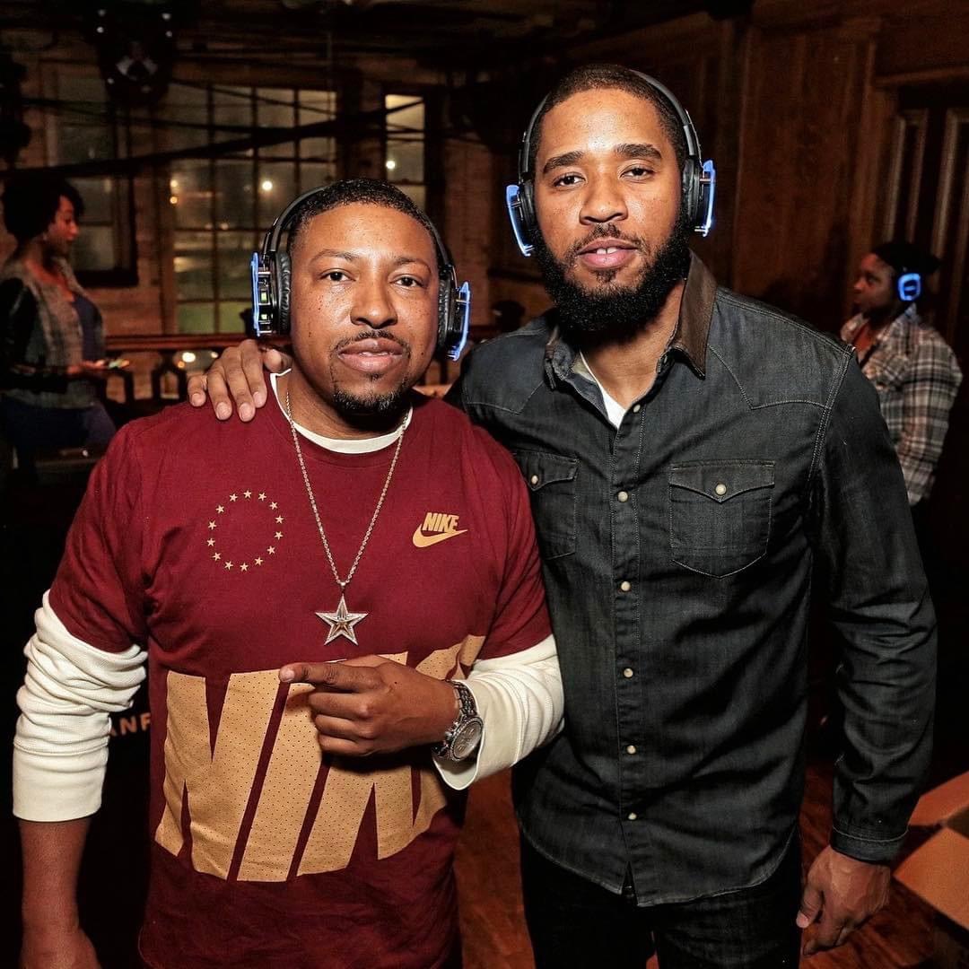 Two men wearing headphones pose together indoors, one in a red Nike shirt and the other in a black shirt, with a wooden background.