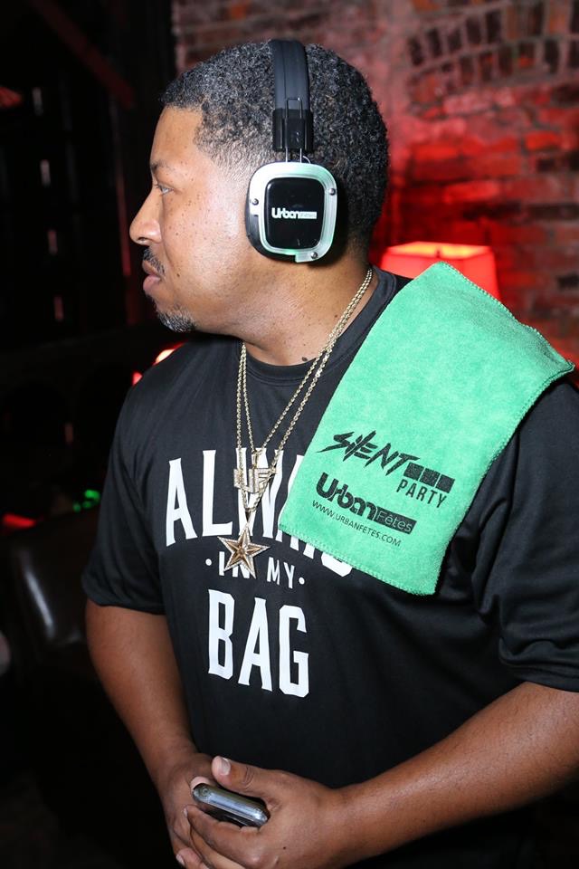 Man wearing headphones and a black shirt with white text, holding a phone, with a green towel over his shoulder, brick wall background.