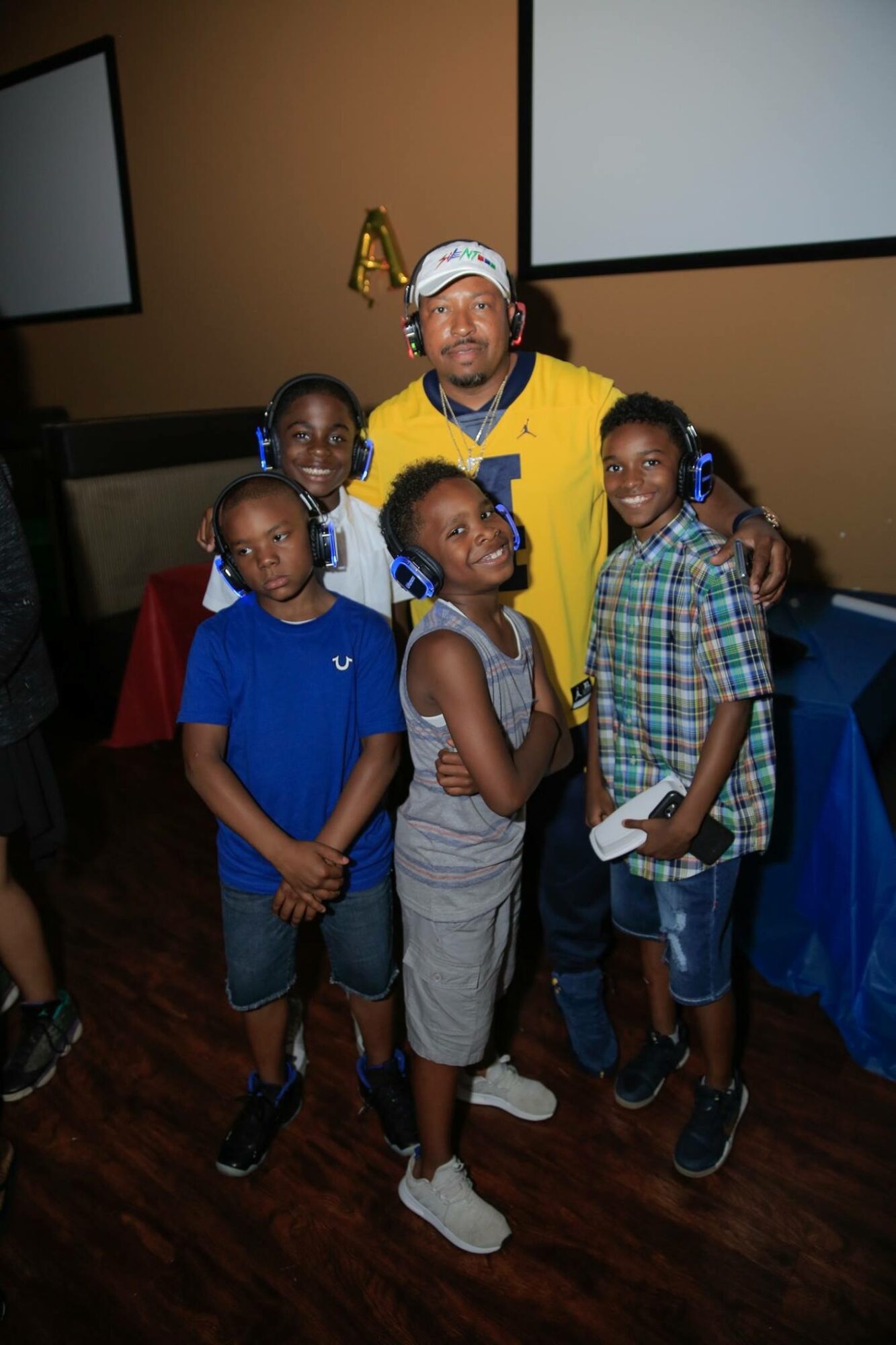 Group of five children and one adult posing indoors, some smiling, with two large screens in the background.