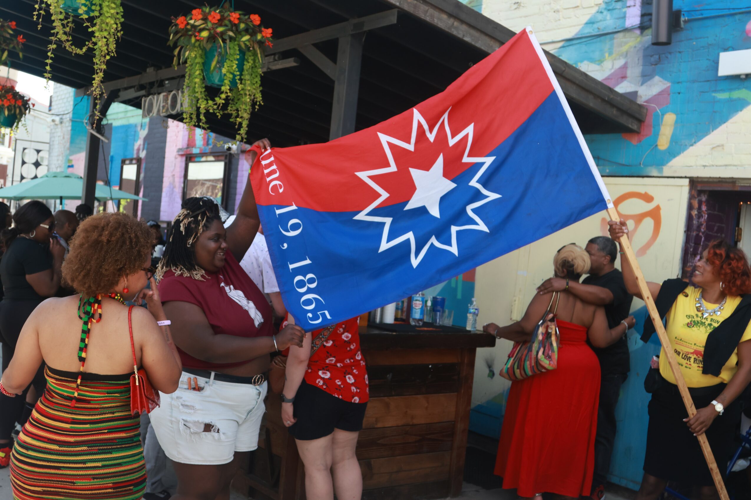 Group of people holding a large flag with a red, white, and blue design, in an outdoor urban setting.
