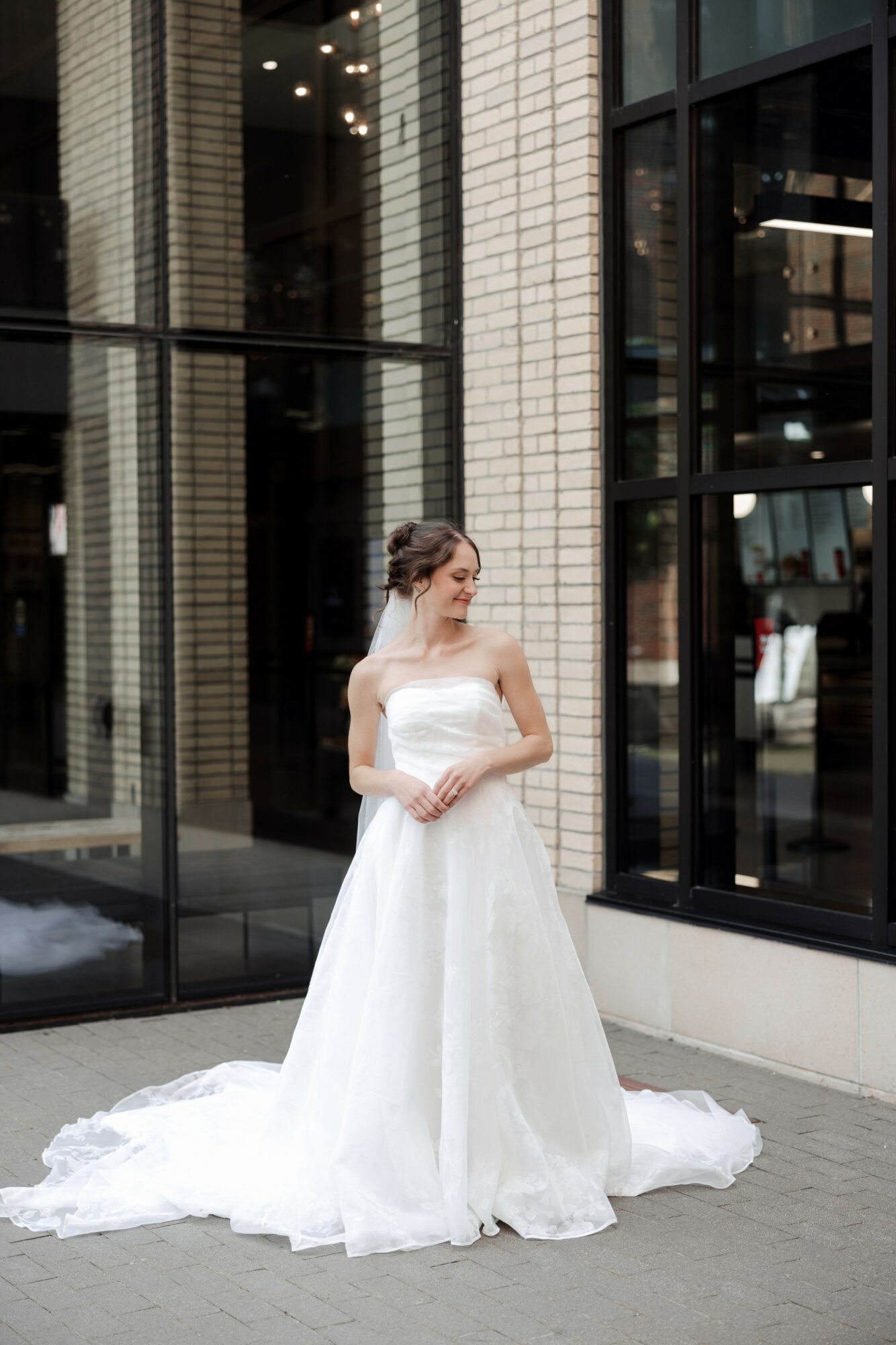 Woman in a white wedding dress standing outside near modern glass building, looking to the side.