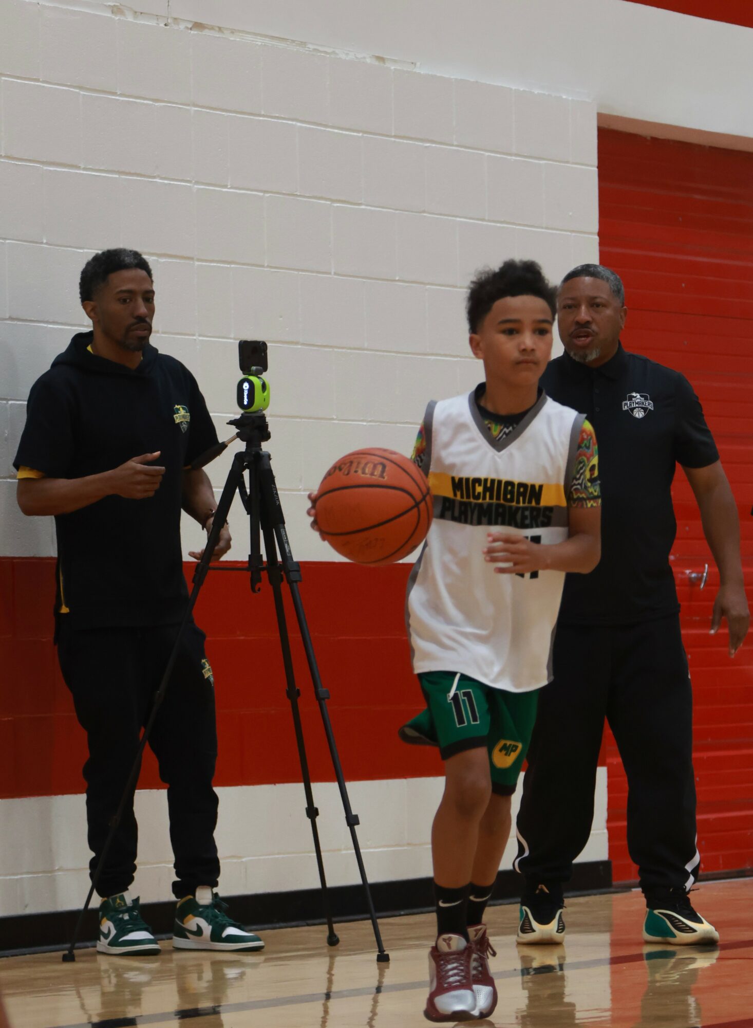Young basketball player holding a ball, with two men standing nearby in a gymnasium.