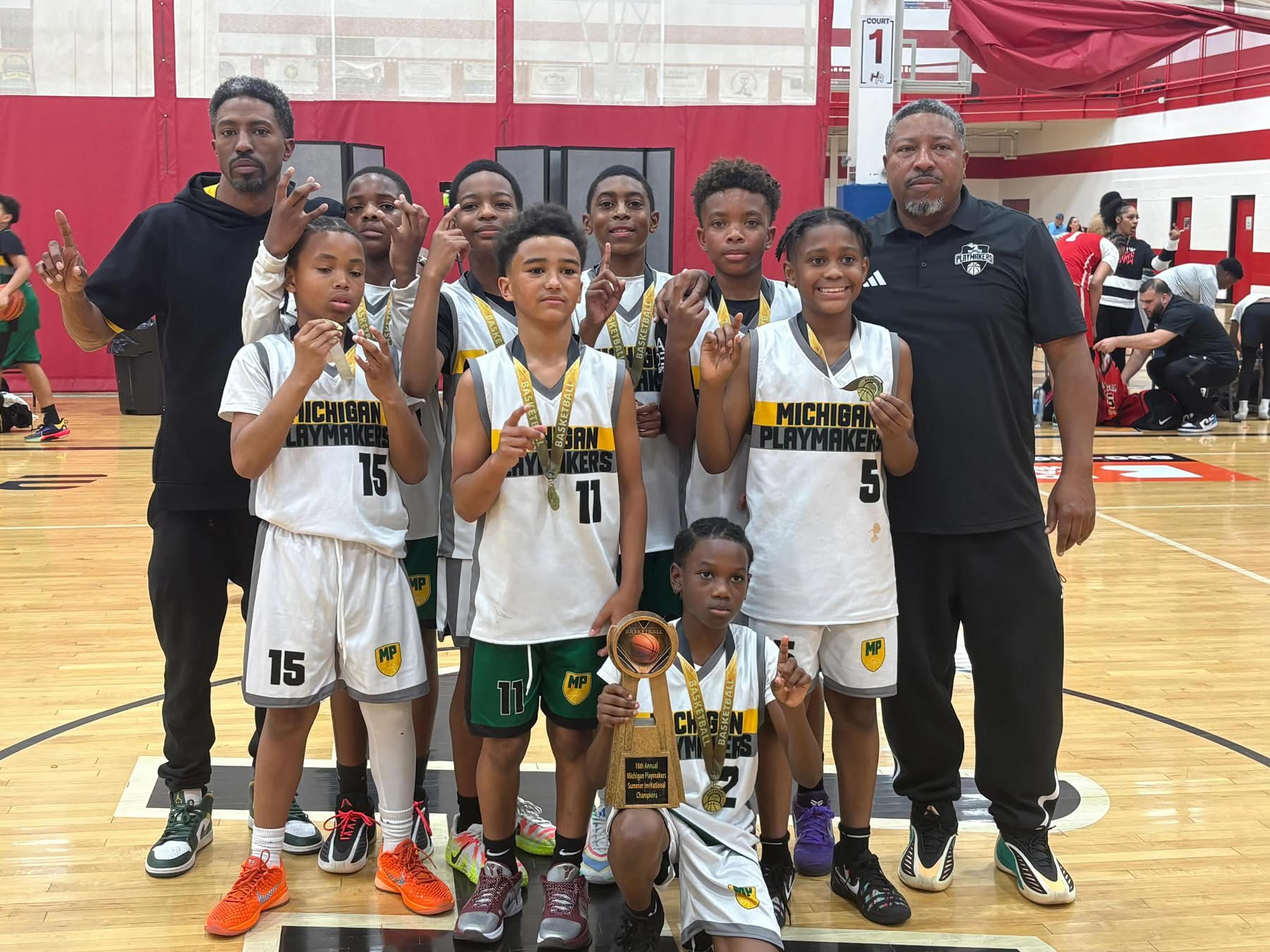 Group of young basketball players and two coaches in a gymnasium, some holding medals and a trophy, celebrating victory.