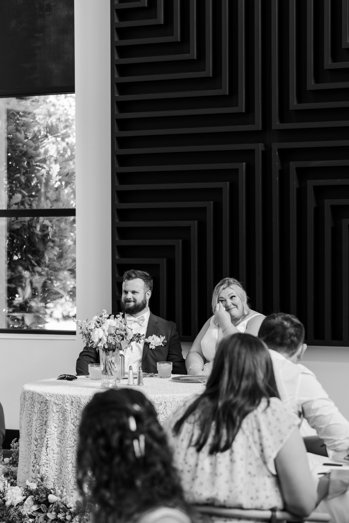 Two people sit at a table with flowers, smiling, in a room with large windows and geometric wall art.