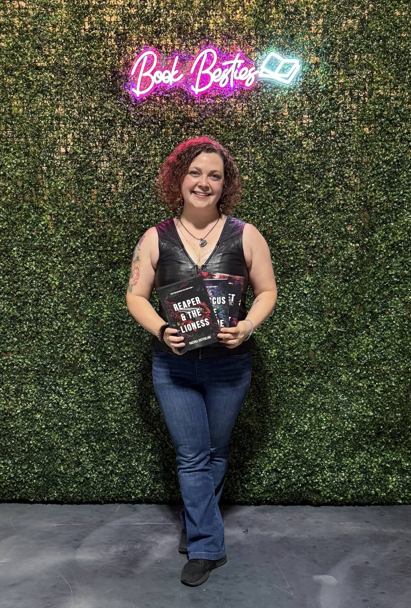 Woman with curly red hair holding books, standing in front of a green wall with neon sign reading 'Book Botique'.