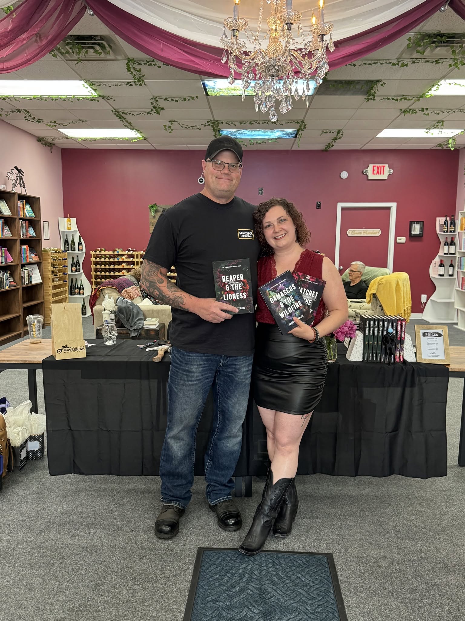 Two people stand in front of a table with books and items in a decorated room, holding books and smiling.