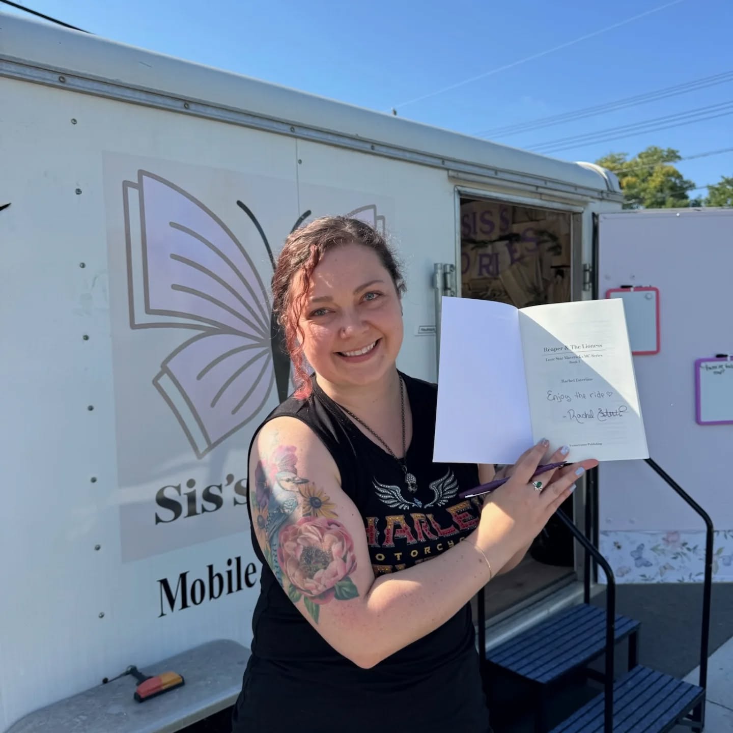 Woman with tattoos holding open book, smiling, standing outside mobile unit with butterfly logo, blue sky in background.